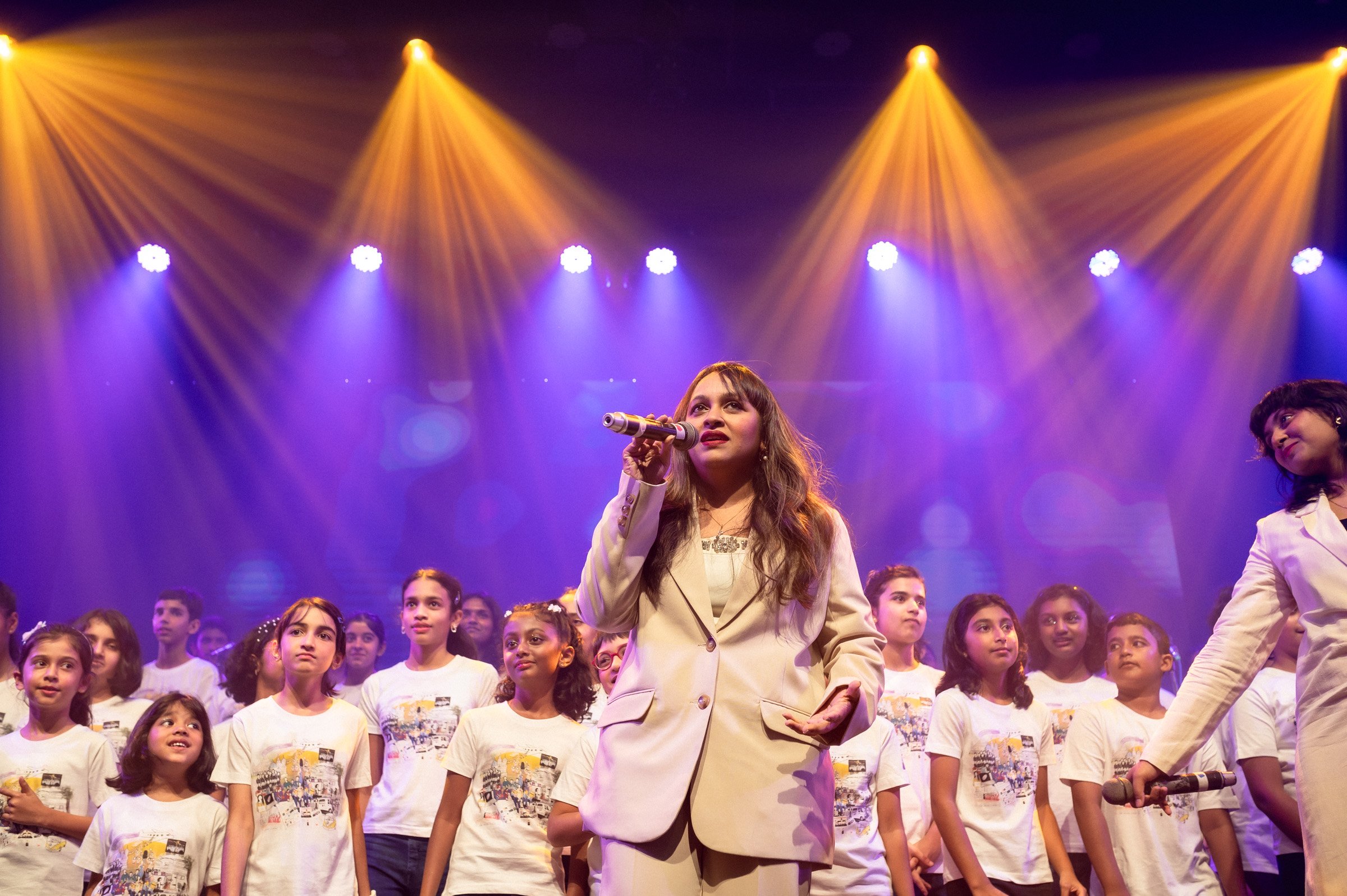 Performer singing on stage with children in white T-shirts, surrounded by colorful stage lights.