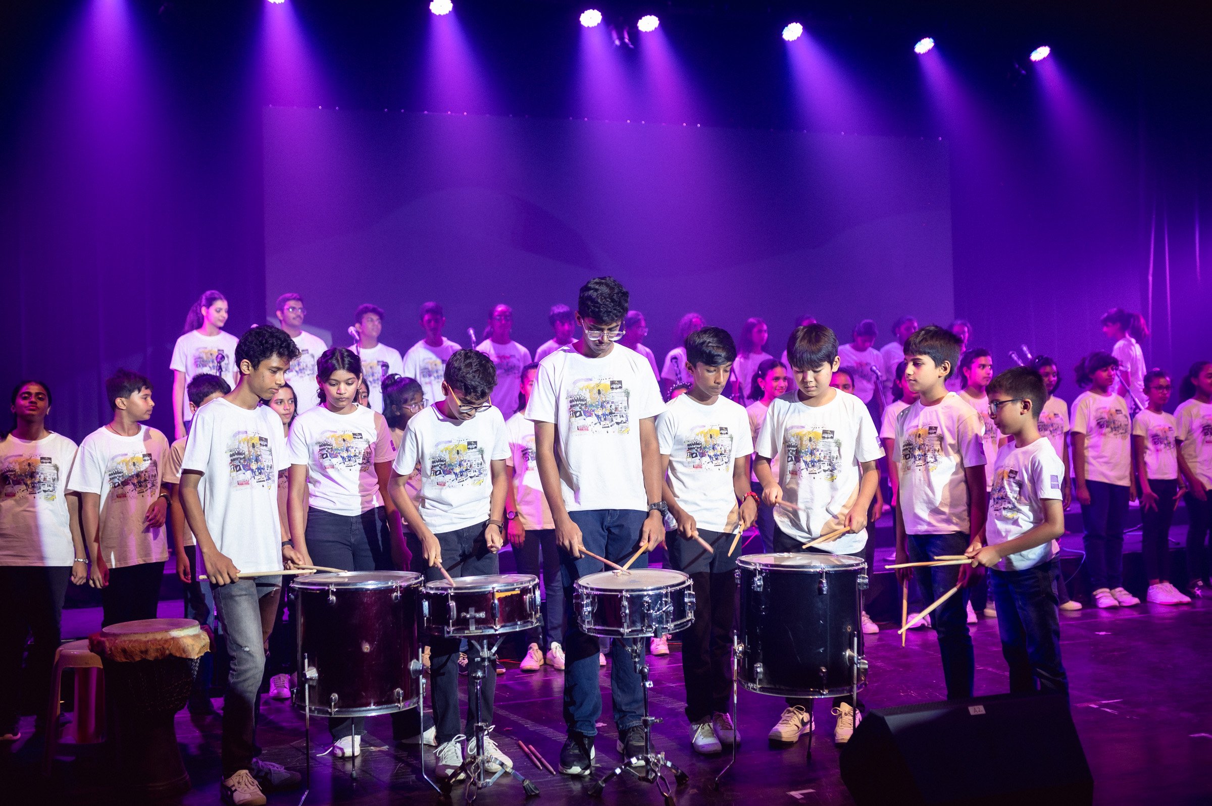 Group of children performing with drums on stage under purple lights, wearing white t-shirts