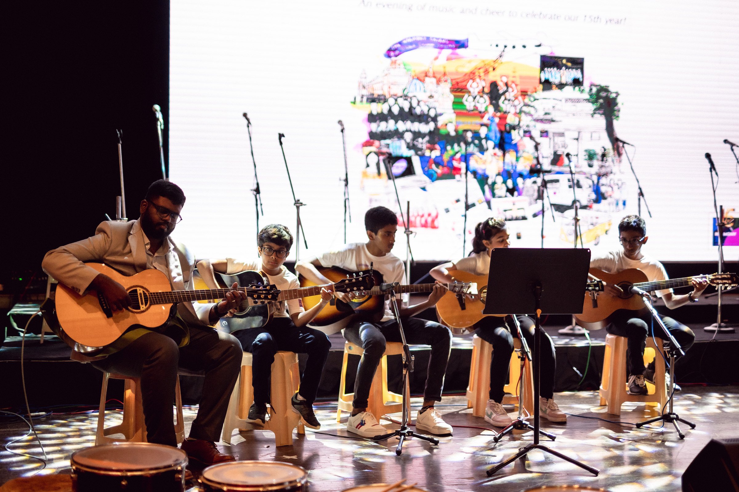Group of musicians playing guitars on stage in front of a collage backdrop, with microphones and a music stand.