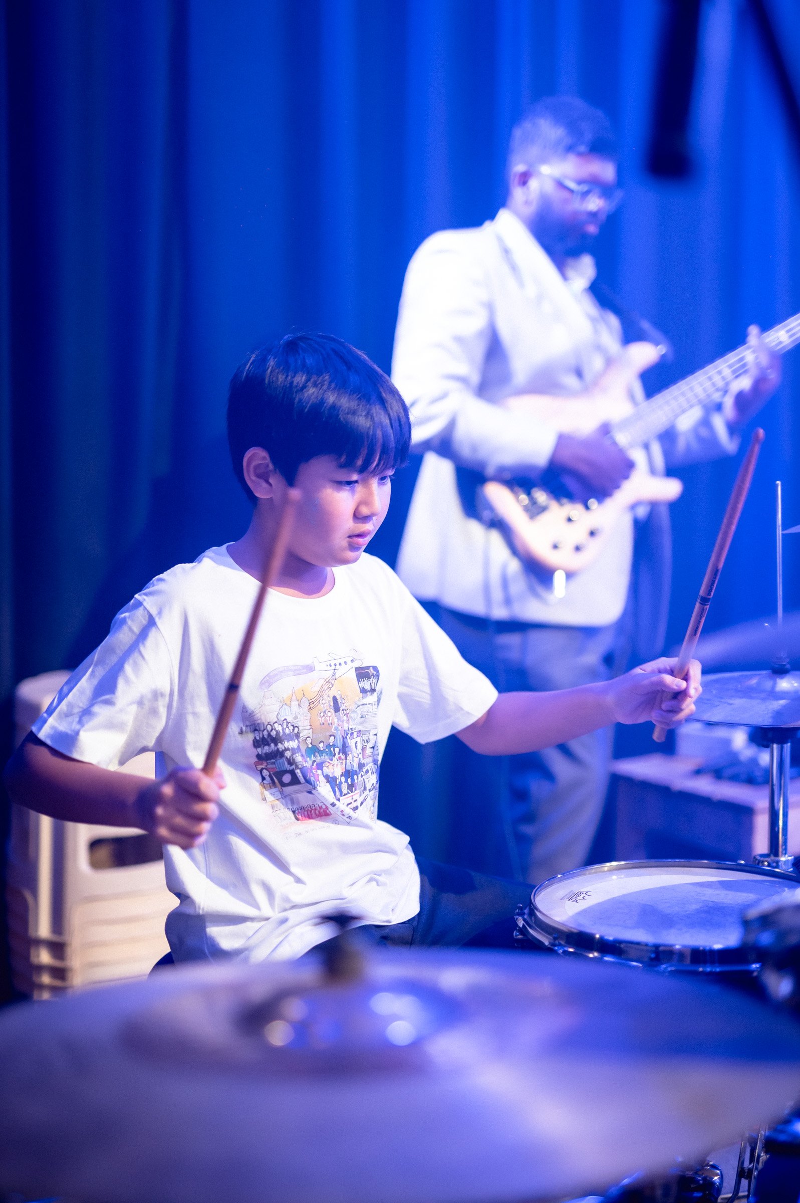 Young boy playing drums on stage with a bassist in the background under blue lighting.