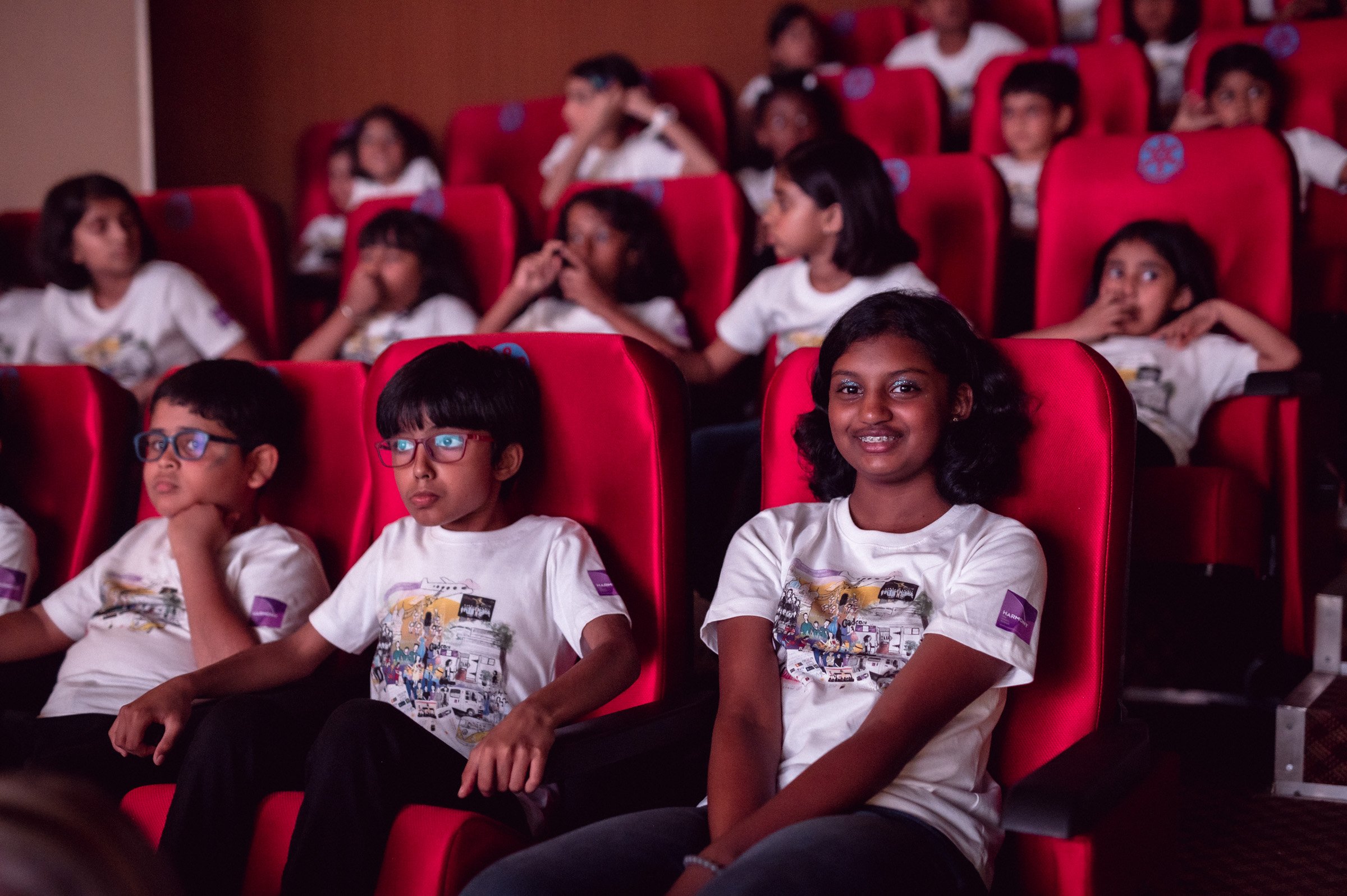 Group of school children sitting in red theater seats, wearing matching white t-shirts, attentively watching a presentation or show.