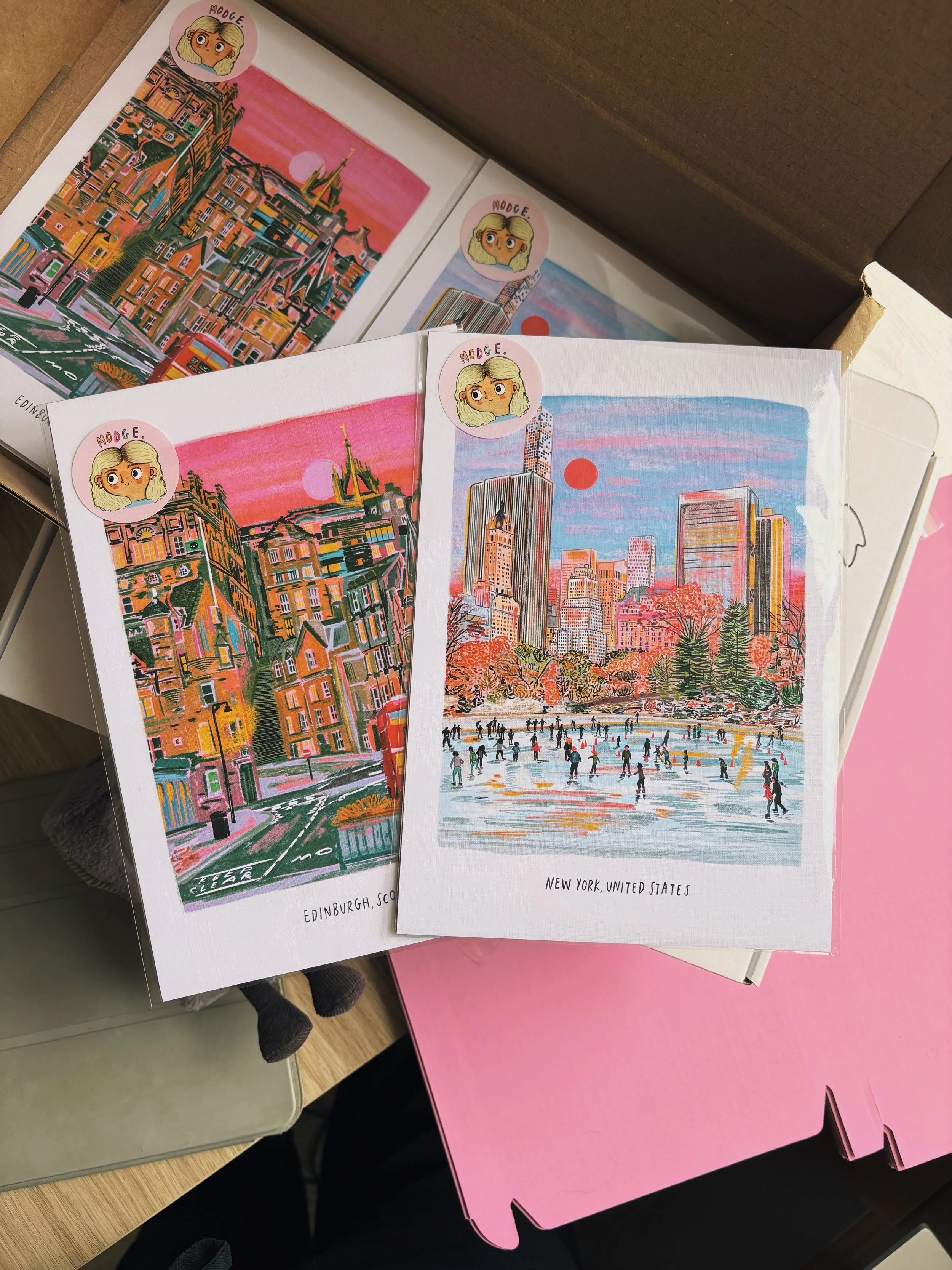 Two illustrated postcards of cityscapes, one of Edinburgh, Scotland, and the other of New York, United States, with colorful, artistic renderings of buildings and landmarks. Both postcards have a sticker with a cartoon girl with blonde hair and the word "MODGE" at the top.