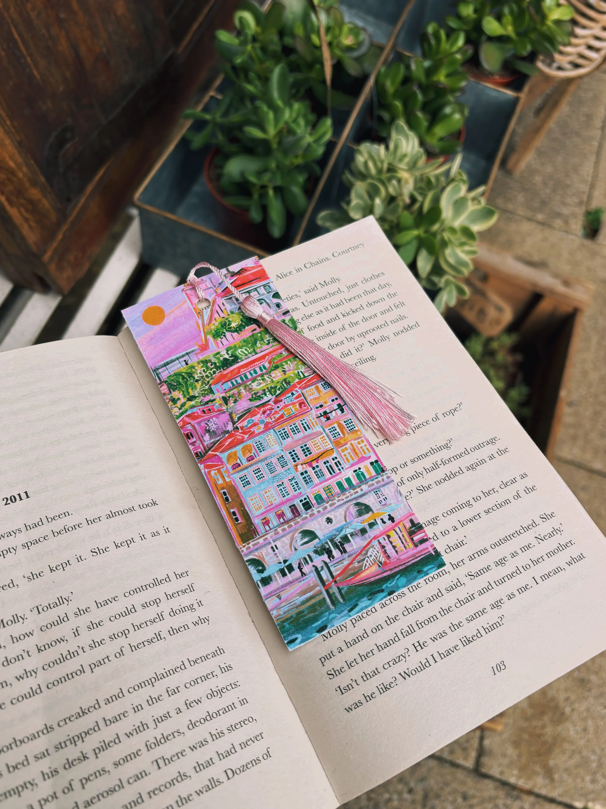 An open book with a colorful bookmark laid across the pages, held near the bottom with a background of potted green plants and a wooden surface.