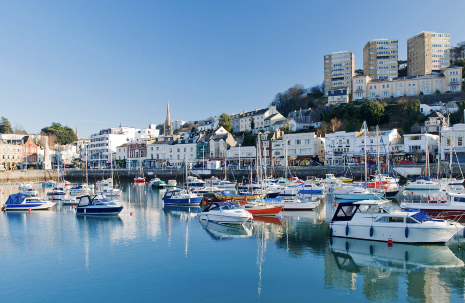 Harbor with numerous sailboats and yachts, waterfront buildings, and hills with high-rise apartments under a clear blue sky.