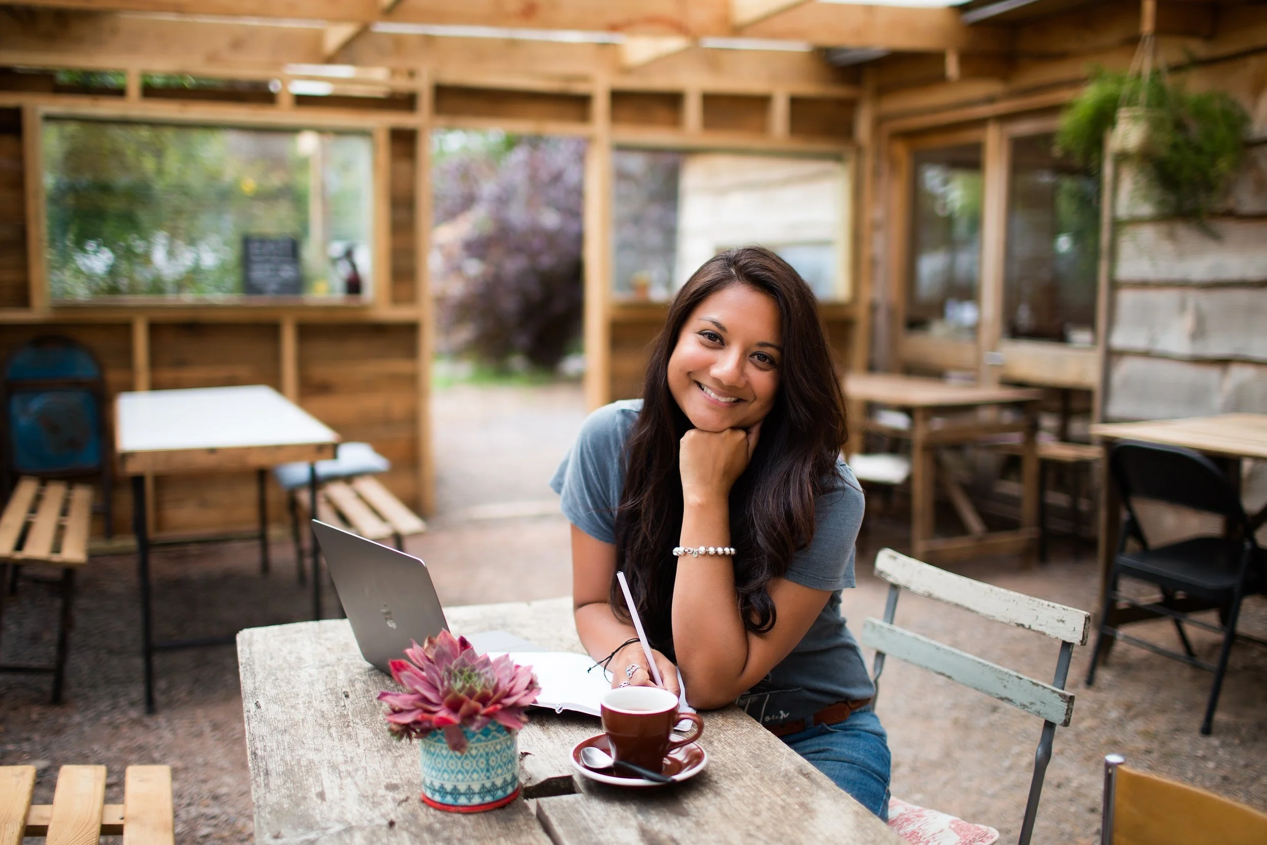 A woman with long dark hair smiling, sitting at a rustic outdoor table with a laptop, a cup of coffee, a notebook, and a succulent plant, in a wooden outdoor cafe.