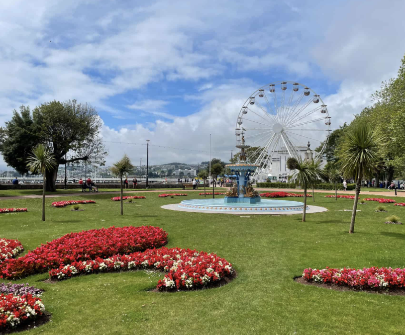 Park with flowers, trees, a fountain, and a large Ferris wheel in the background under partly cloudy sky.