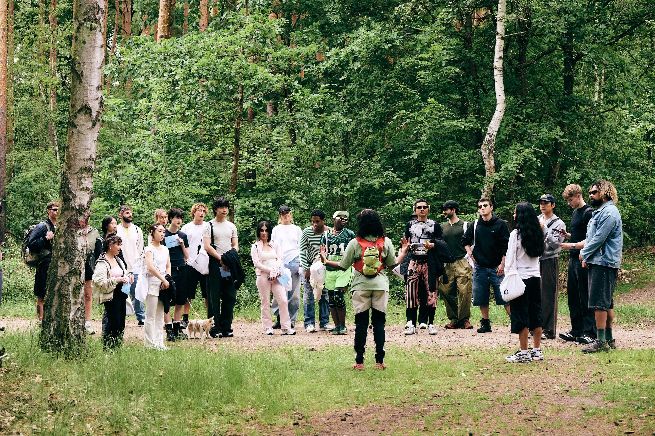 Group of diverse people gathered outdoors in a forest setting, listening to a woman in a green shirt with a red backpack who appears to be leading a tour or giving a talk.