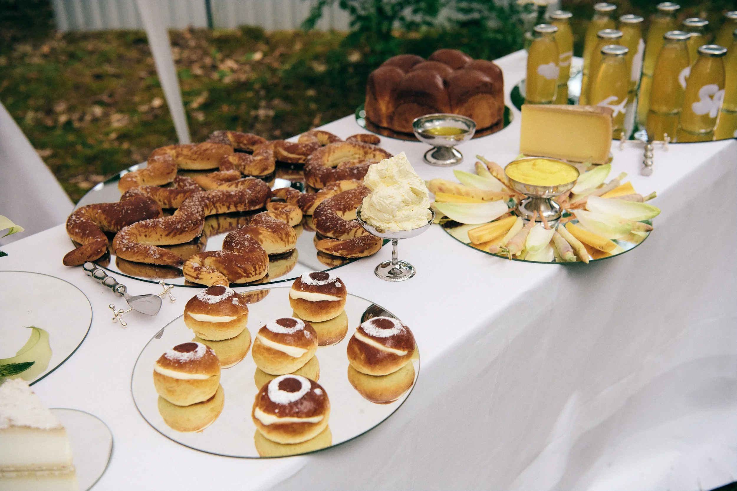 Sweet treats and cheeses on a white tablecloth, including cream-filled cookies, bagels with sugar, a loaf of cake, and a cheese wheel, with some fruit slices and bottled drinks in the background.