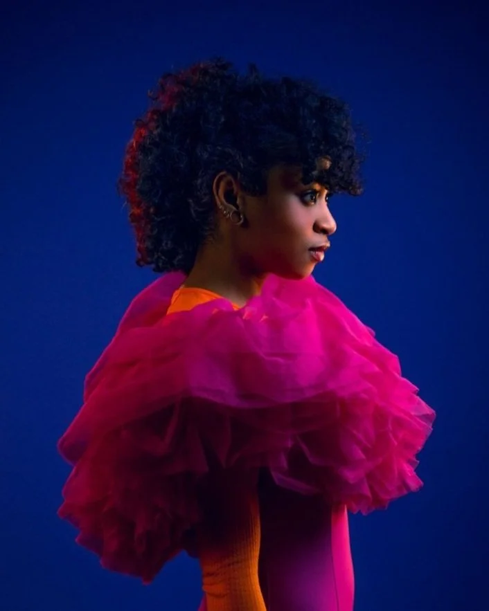 Portrait of Oumou Aidara wearing a vivid pink garment, looking left against a blue backdrop. Berlin-based artist, writer, and athlete.