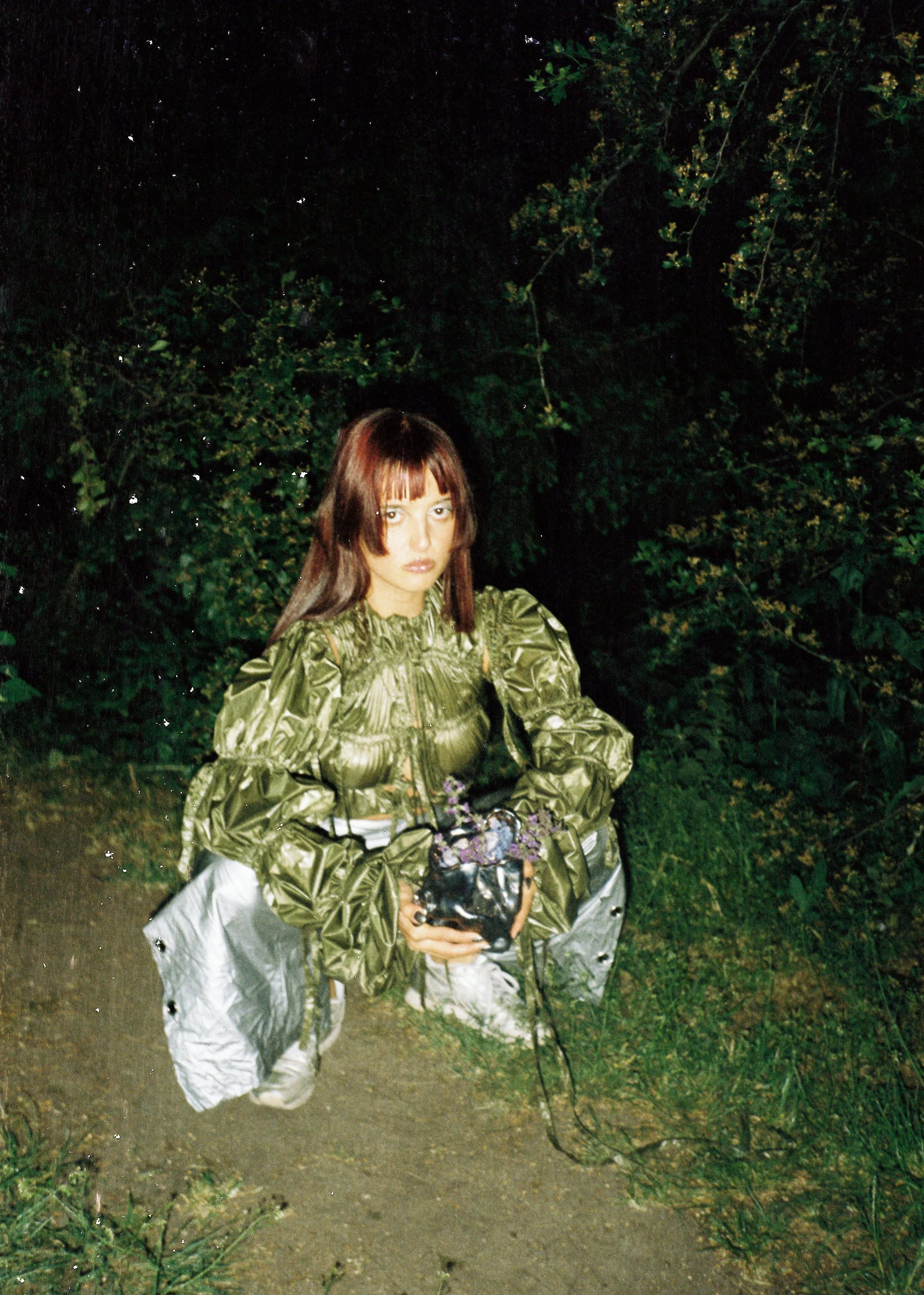A woman with brown hair and bangs kneeling on a dirt path in a wooded area at night, wearing a shiny green jacket and silver pants, holding a potted plant with purple flowers.