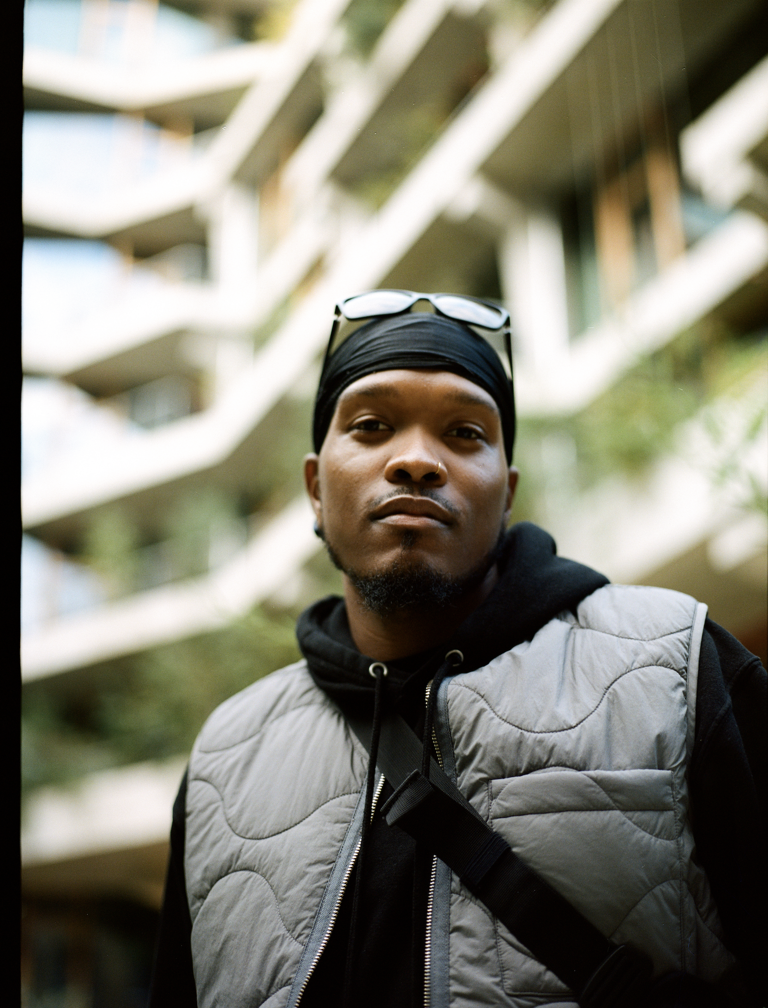 Close-up of a young man with wearing a black headscarf, sunglasses on his head, a nose ring, and a gray quilted vest over a black hoodie, standing outdoors with blurred apartment buildings and greenery in the background.