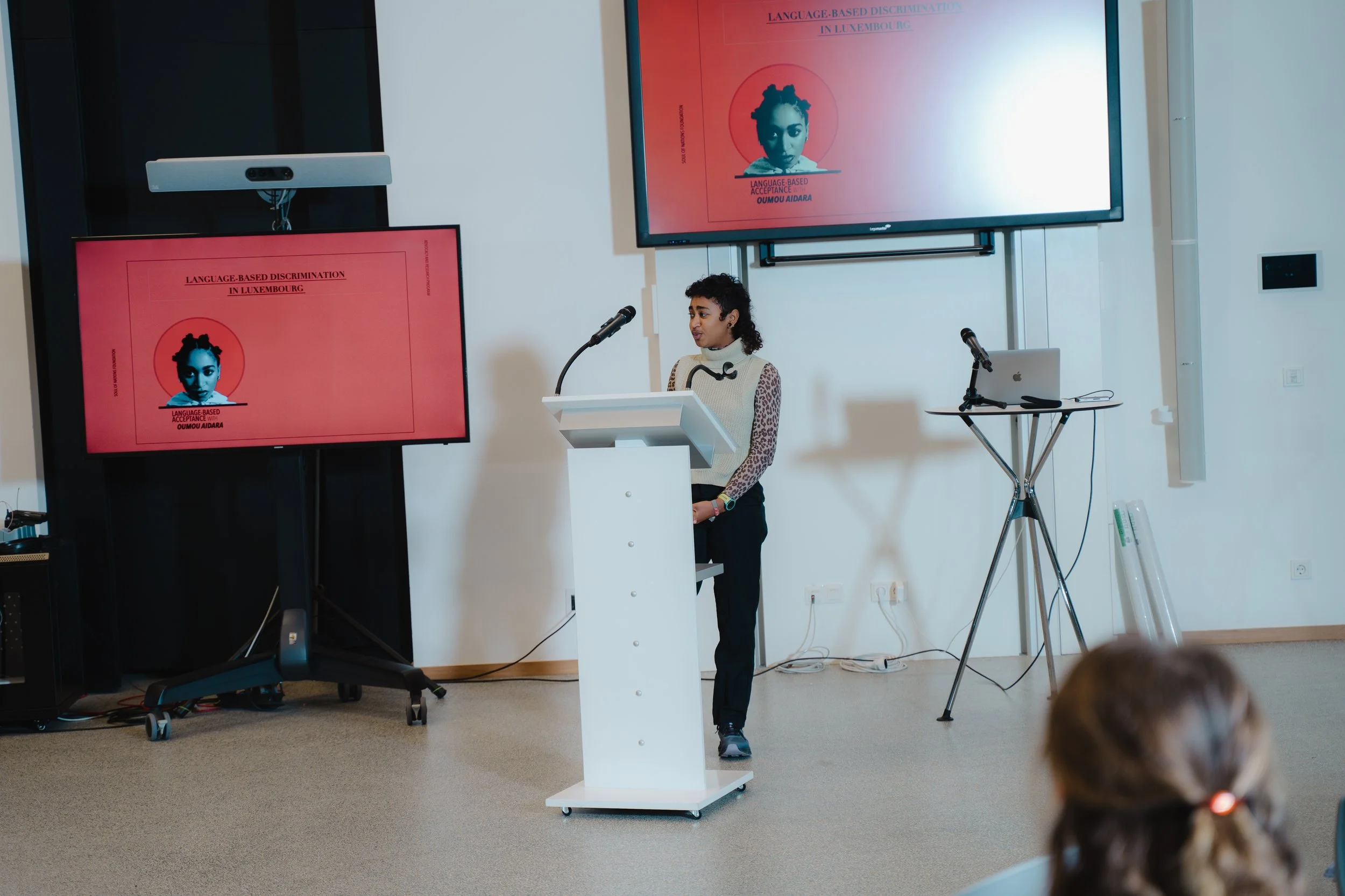 A woman giving a presentation at a conference, standing behind a white podium with a microphone. Two screens display the presentation titled 'Language-Based Discrimination in Luxembourg' with an image of a woman's face.