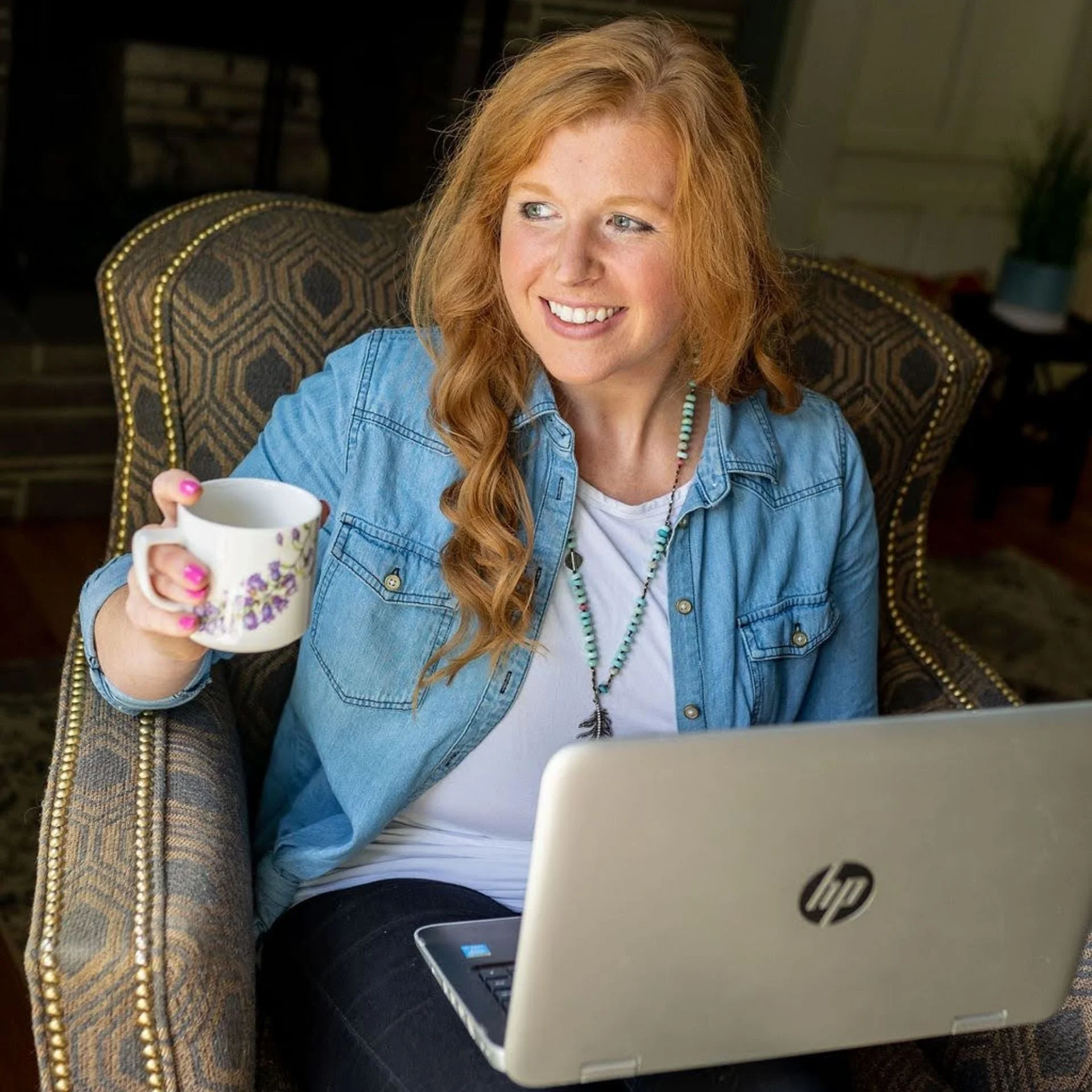 Maggie, a professional home organizer in Rochester, smiling while holding a coffee mug and working on a laptop in a comfortable armchair.