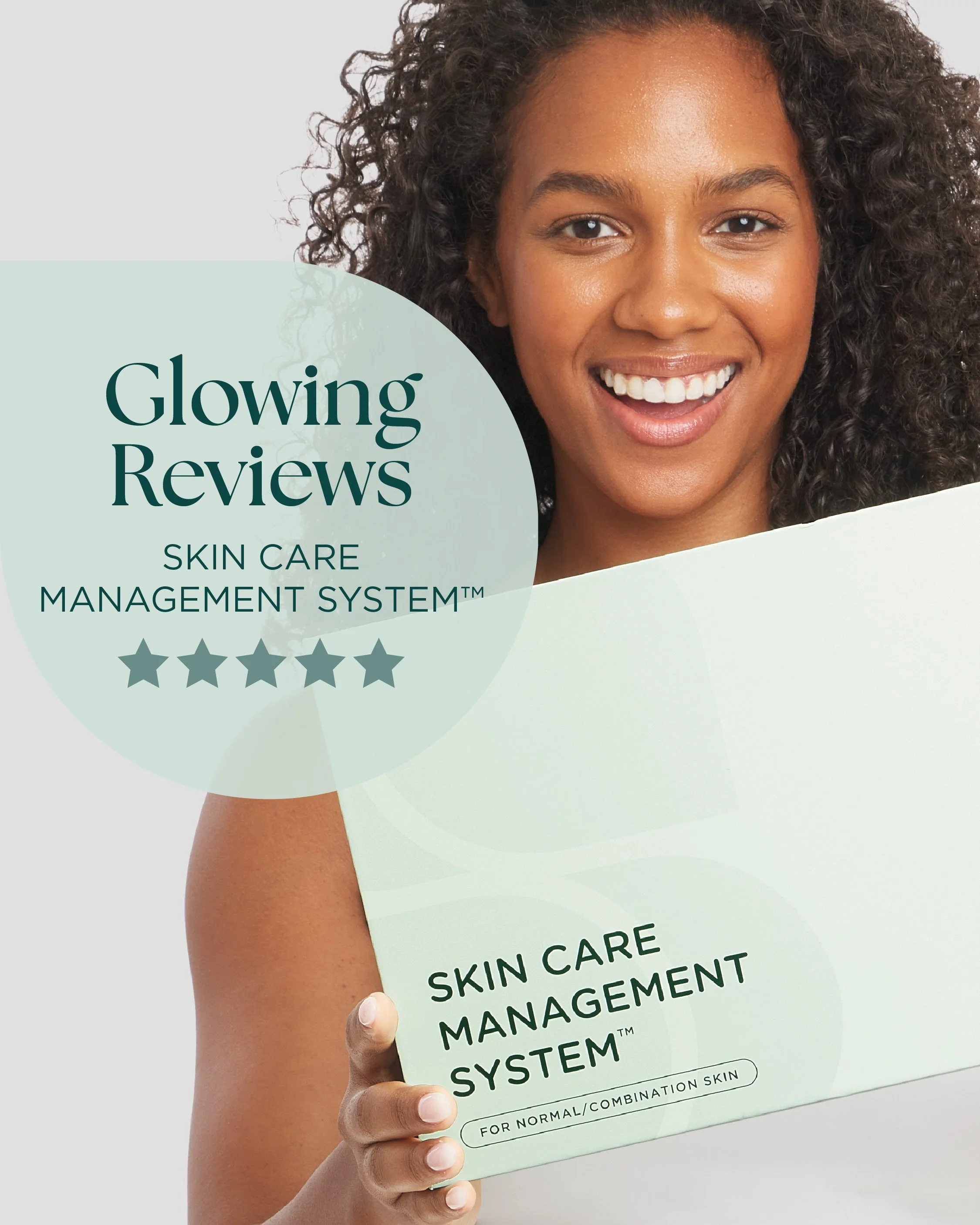 A woman with curly hair holding a skincare management system box with a big smile, showing her teeth, against a plain light background.