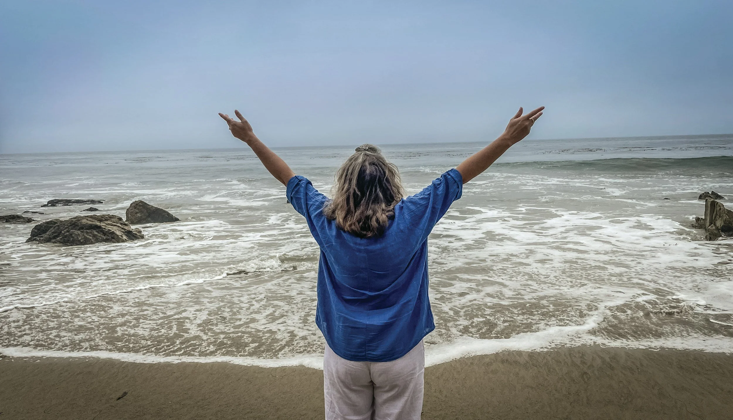 midlife woman in long sleever blue shirt and white pantsstanding in front of the beach with arms outstretched
