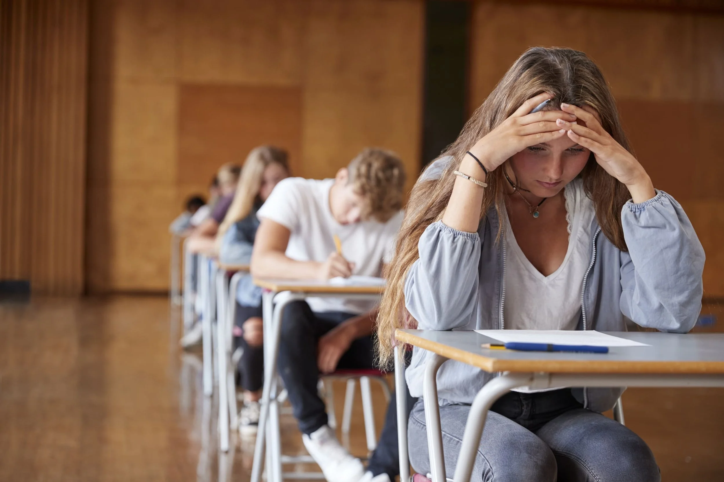 Anxious teenage student sitting at a desk during an exam while classmates work in the background, representing academic pressure and stress during the transition to college.