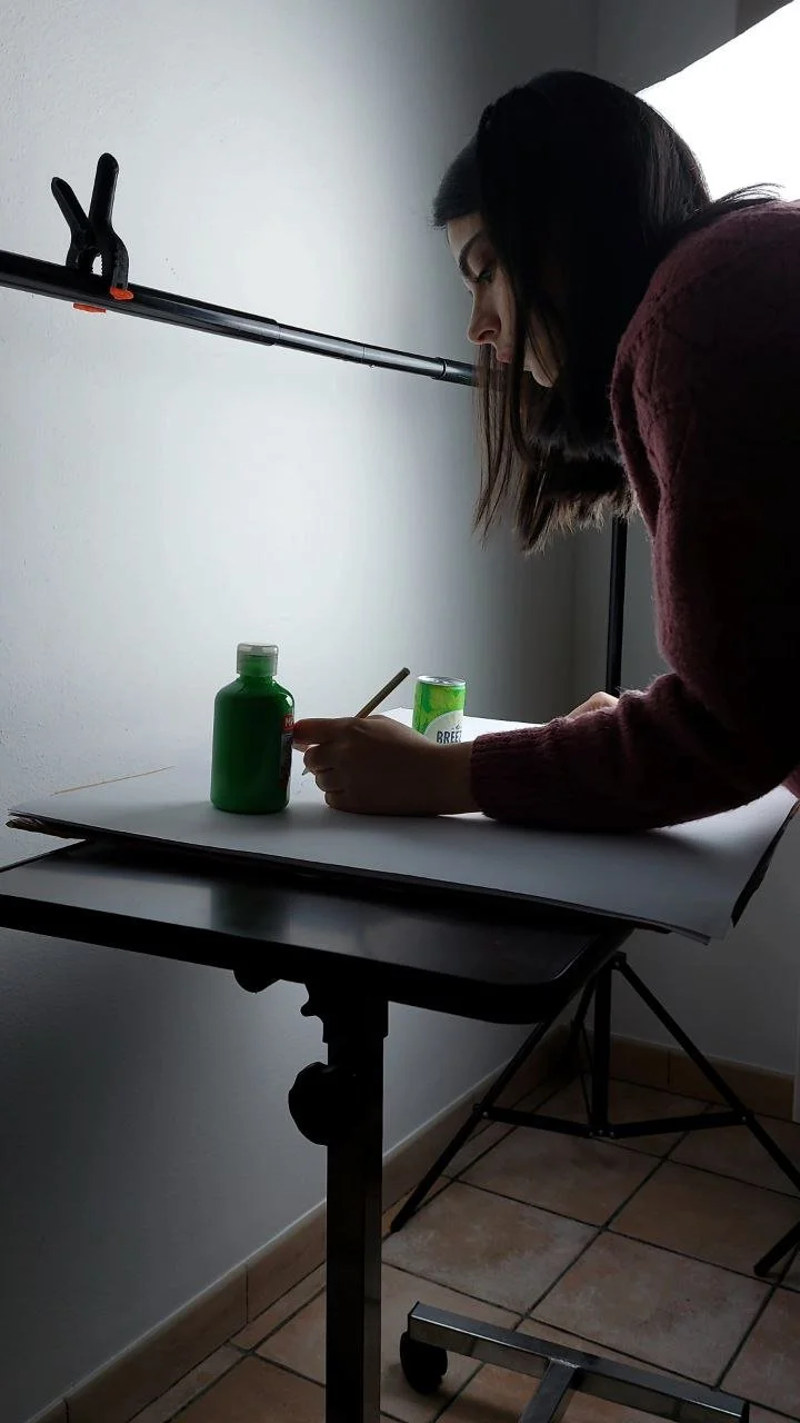 Ludovica Floris leaning over a table, working on a project with a Breezer, and a pencil, in a room with a white wall and tiled floor.
