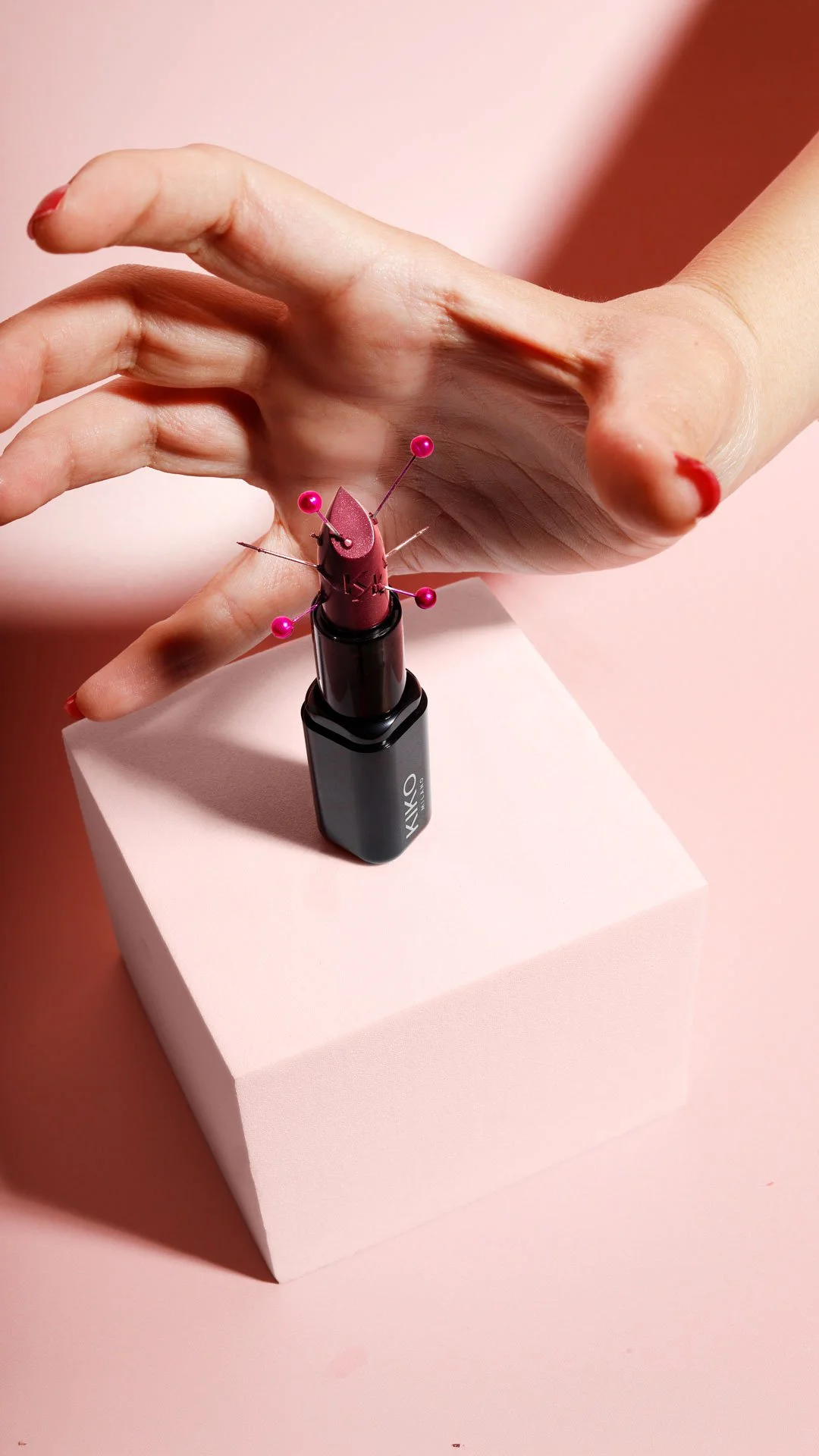 Editorial beauty product photography — a hand with red nails reaching toward a Kiko Milano lipstick pierced with hot pink sewing pins, on a white cube against a soft pink background with hard light shadows. Art direction and photography by Ludovica F