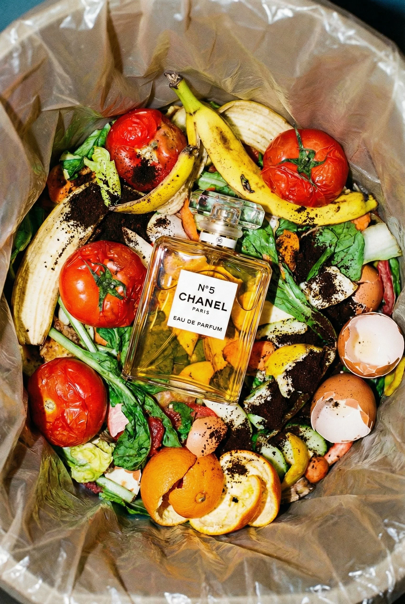 A collection of rotting fruits and vegetables inside a trash bag, with a bottle of Chanel No. 5 perfume placed on top.