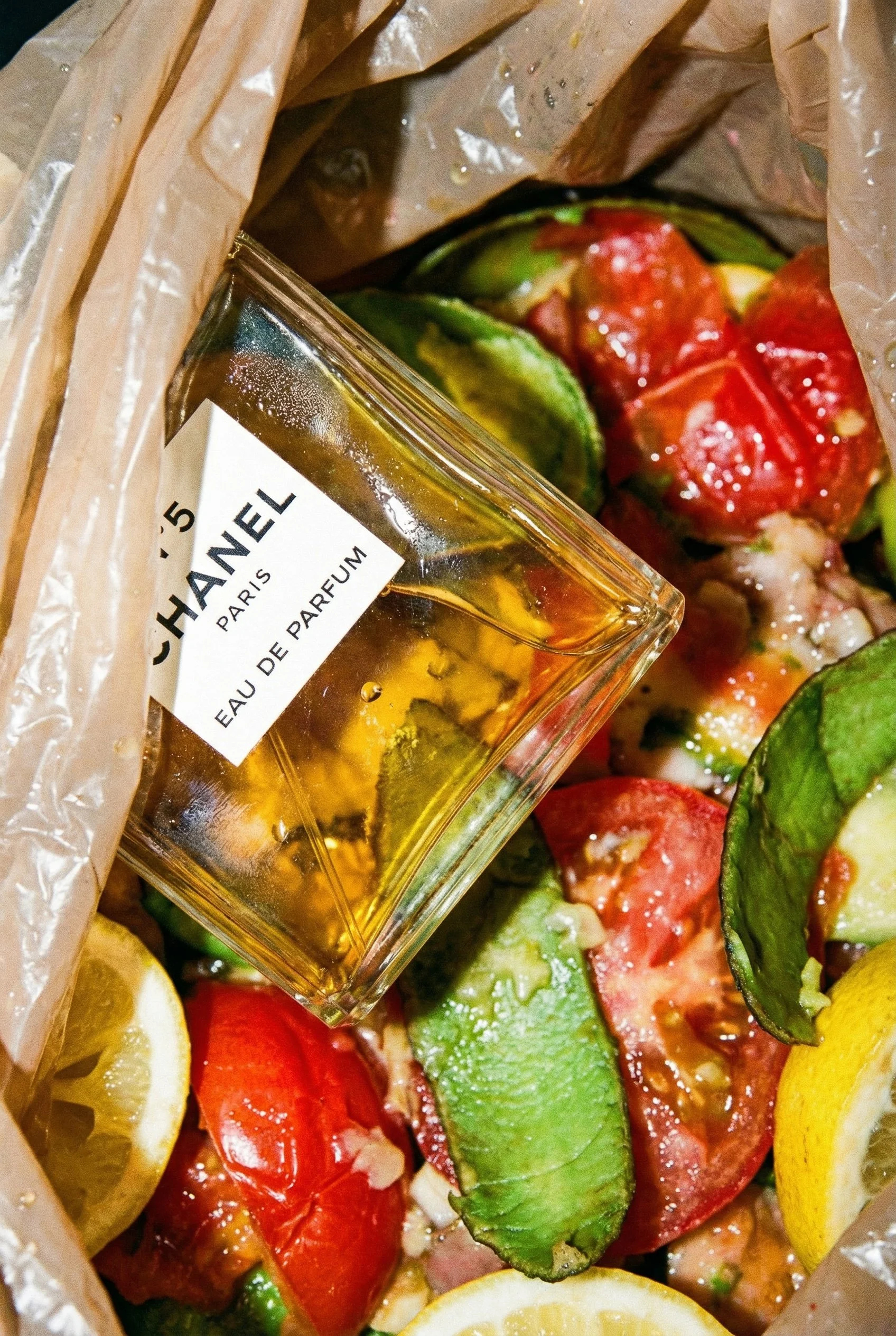 A bag containing red and green tomatoes, lemon slices, and a bottle of Chanel No. 5 perfume amidst the tomatoes.