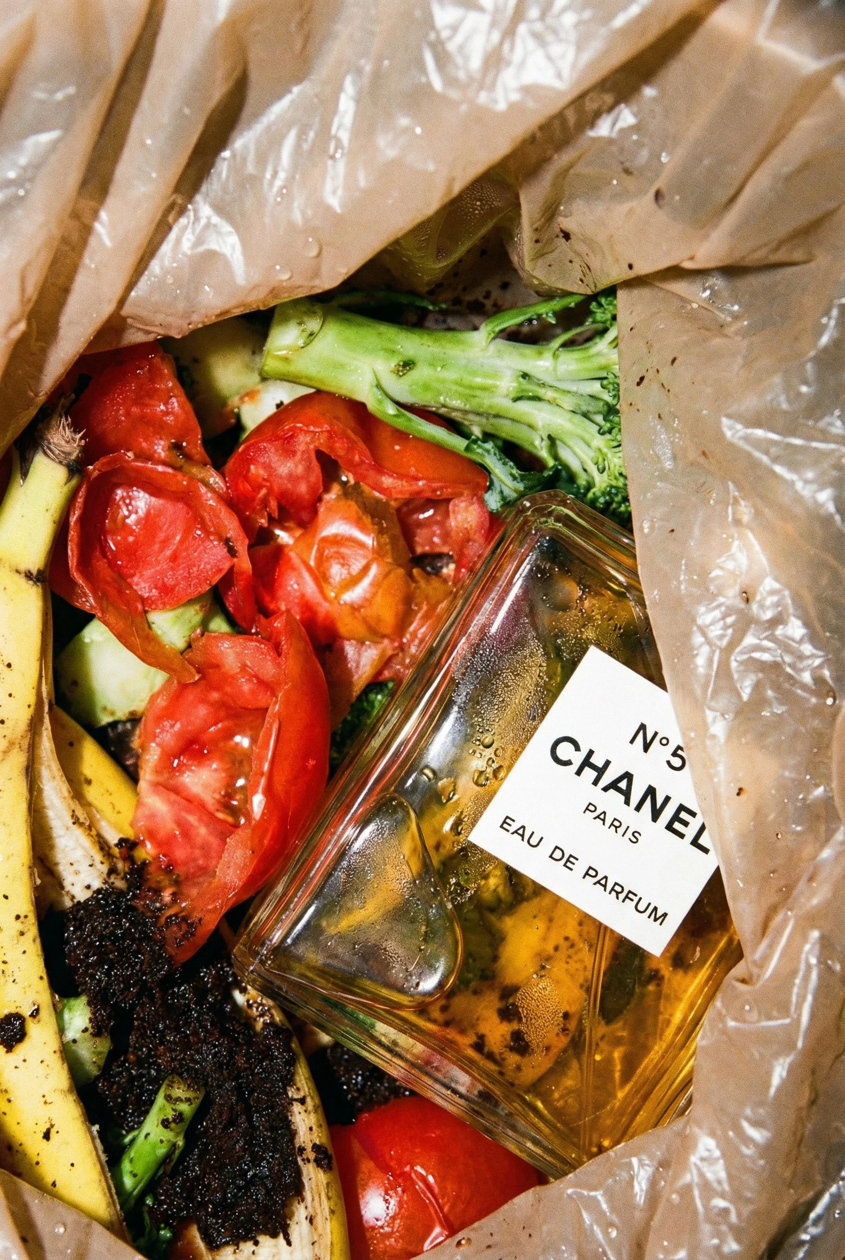 A collection of vegetables including tomatoes, broccoli, and bananas surrounding a bottle of Chanel Eau de Parfum inside a paper bag.