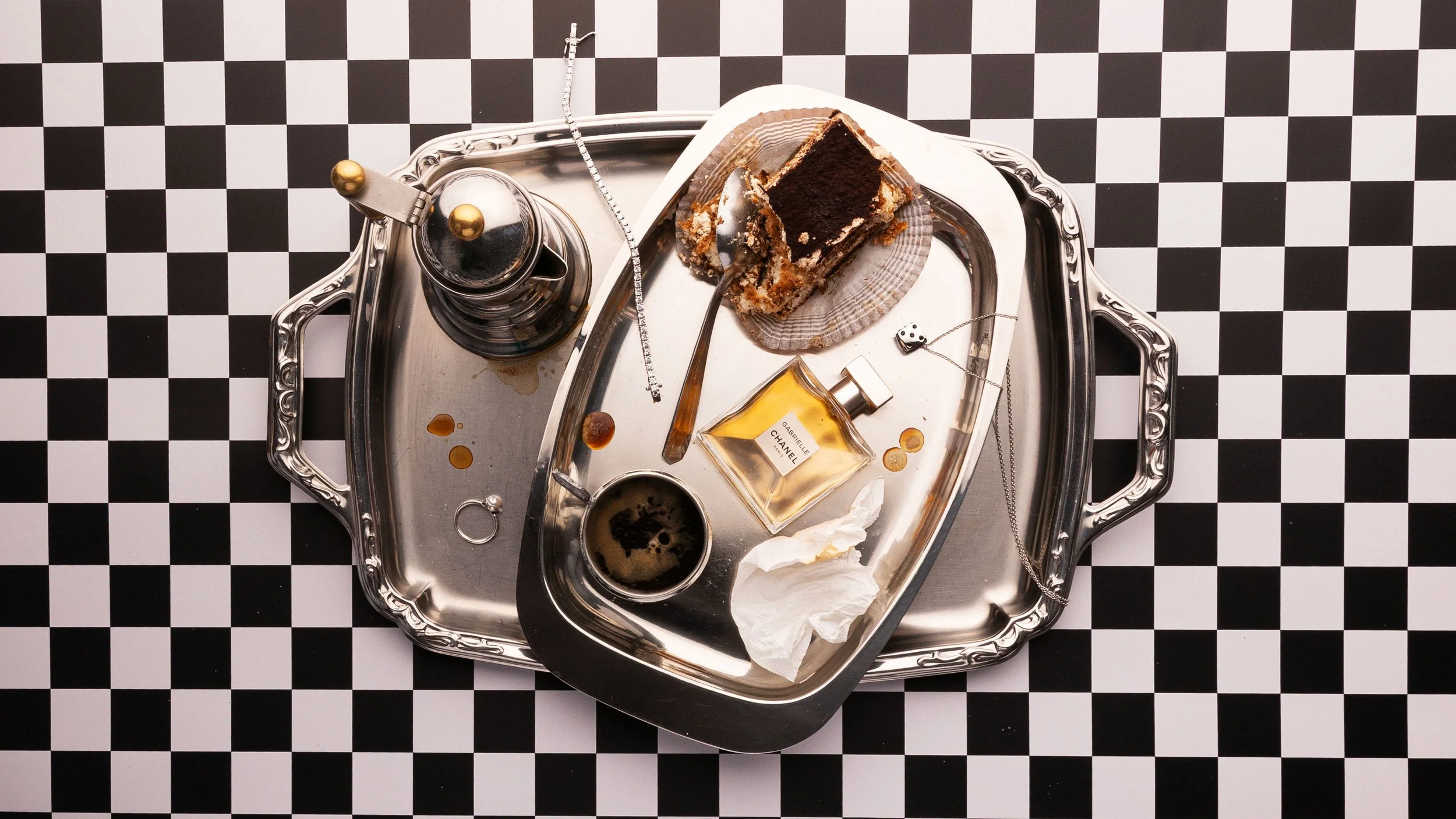 Flat lay of a silver tray with Gabrielle Chanel perfume bottle, tiramisu slice, espresso, and jewelry on a black and white checkered surface.