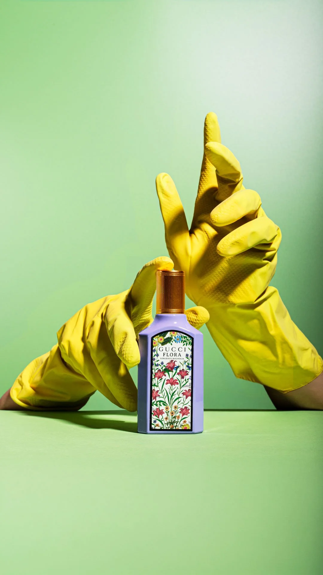 A person wearing yellow rubber gloves holding a Gucci Flora perfume bottle with a floral label, set against a green background.