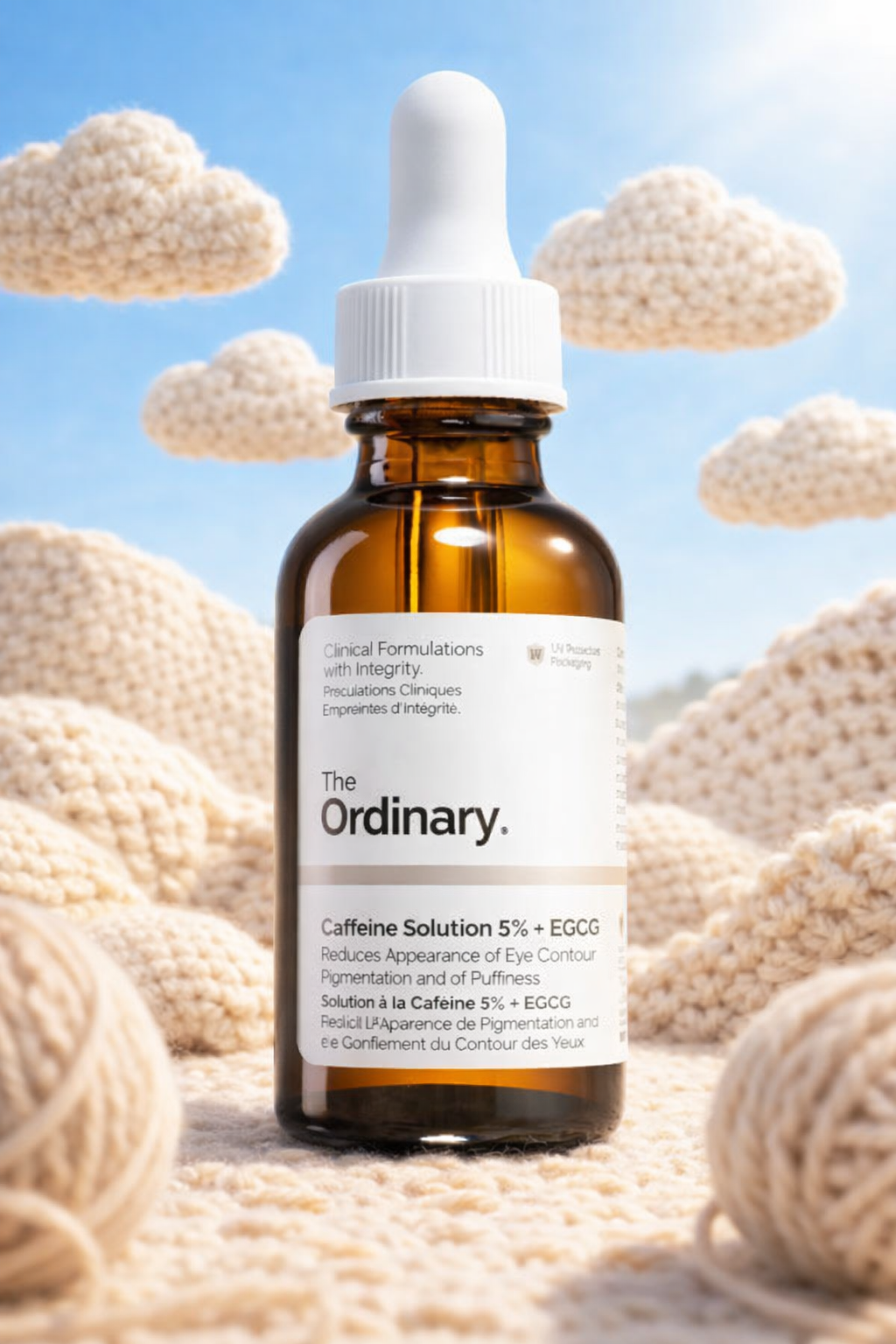 A brown glass bottle of The Ordinary Caffeine Solution 5% + EGCG with a white dropper cap, set against a background of cream-colored knitted clouds and yarn balls, under a blue sky.