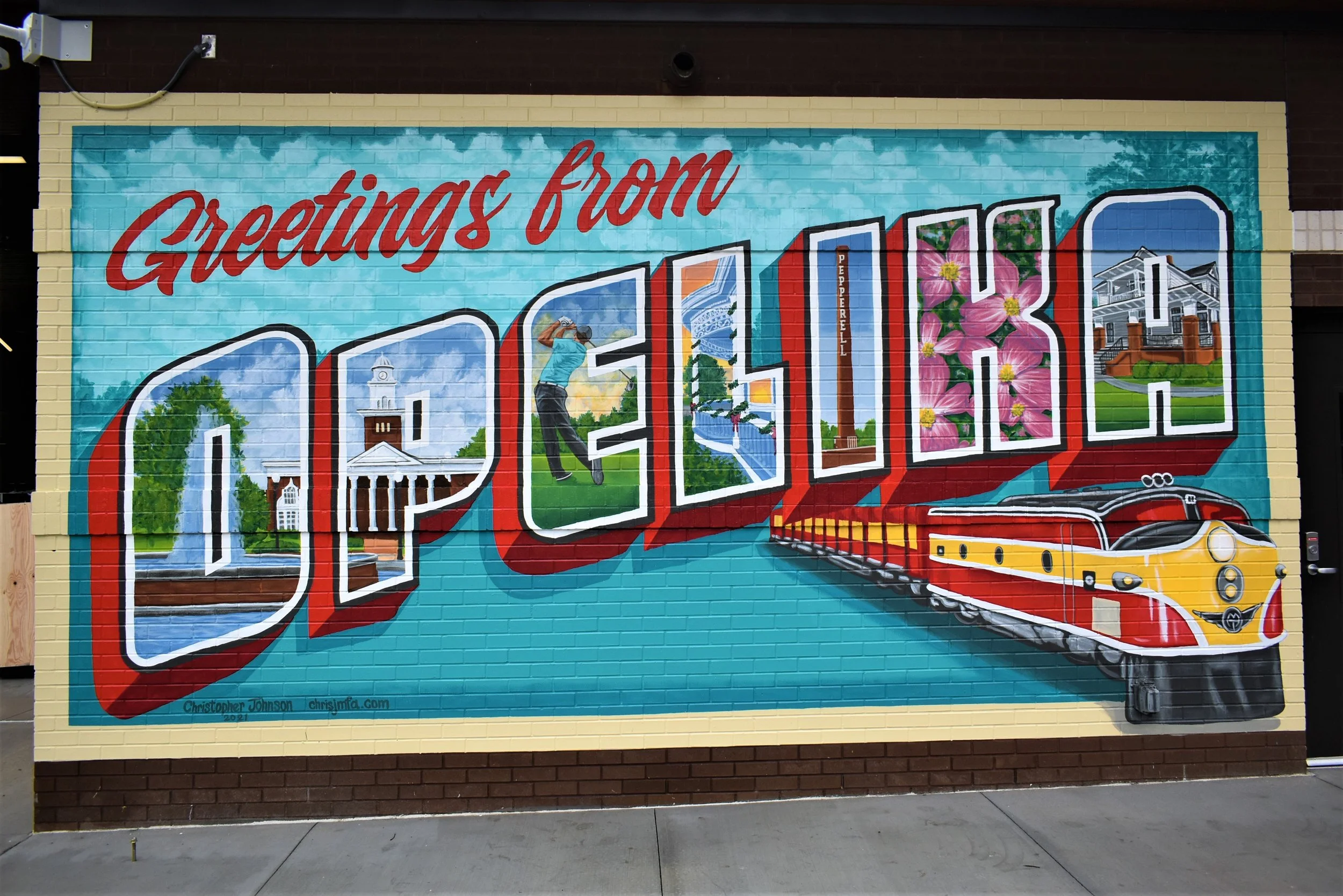 Mural that reads "Greetings from Opelika" with a train, golf player, flowers, and other important landmarks.