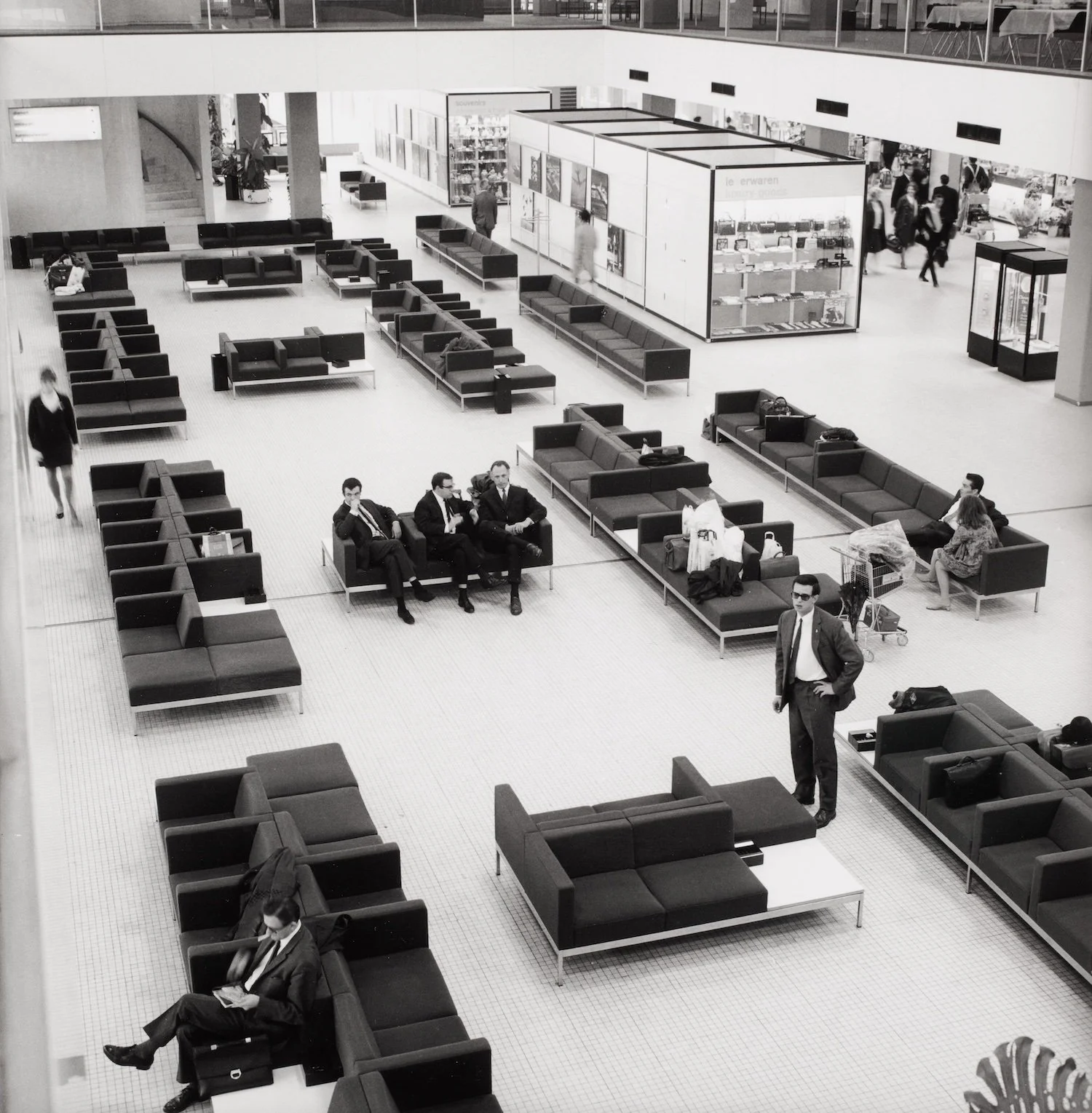 Black and white photo of an airport waiting area with black couches, some occupied by passengers, some empty, and a few standing or walking around. A glass display case and people walking past in the background.