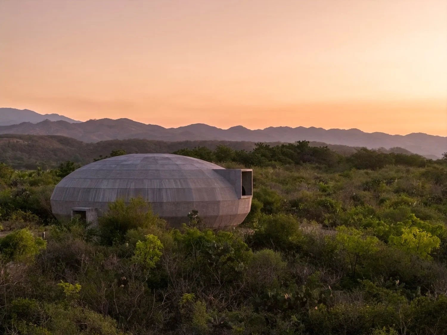 Round, domed concrete building surrounded by green bushes in a desert landscape at sunset with mountains in the background.