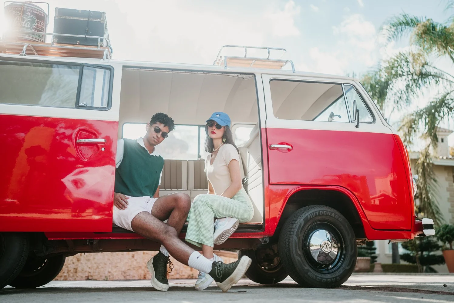 Two young adults sitting inside a red and white vintage van parked on a street, with trees and buildings in the background.