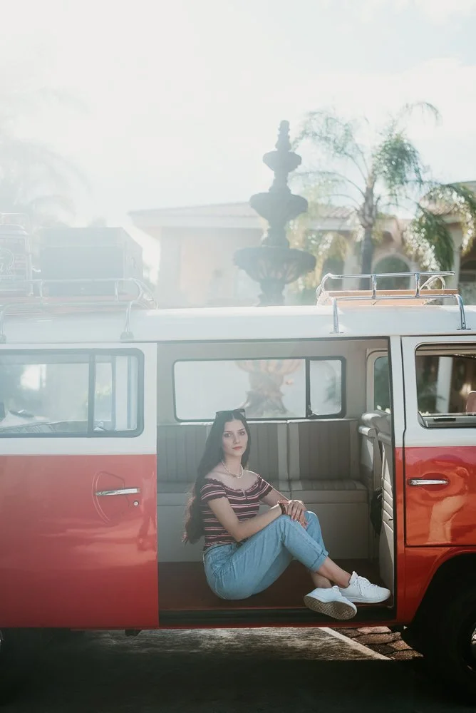 A young woman with long dark hair, wearing a striped top, jeans, and white sneakers, sitting inside a vintage red and white van, with a fountain and trees in the background.