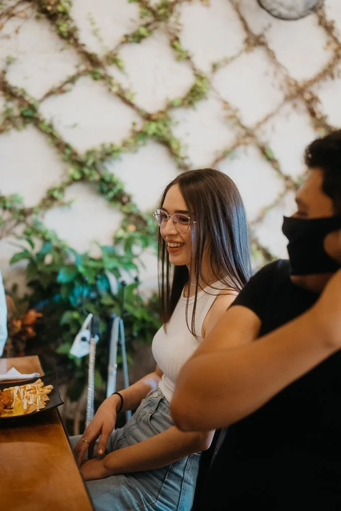 A young woman with long brown hair, glasses, and a white tank top is smiling while sitting at a restaurant table, with a person wearing a black shirt and face mask next to her. There are plants and a textured wall in the background.