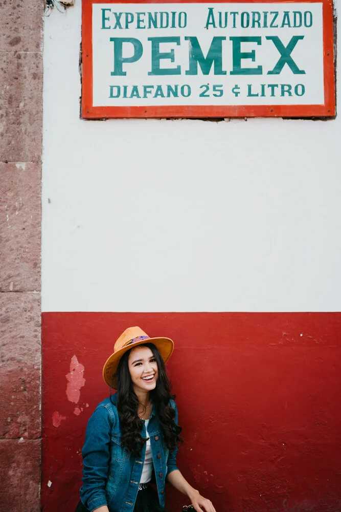 A young woman with long dark hair wearing a straw hat and denim jacket, smiling and standing in front of a red and white wall with a sign that says "Expedio Autorizado PEMEX Diafano 25 & Litro."
