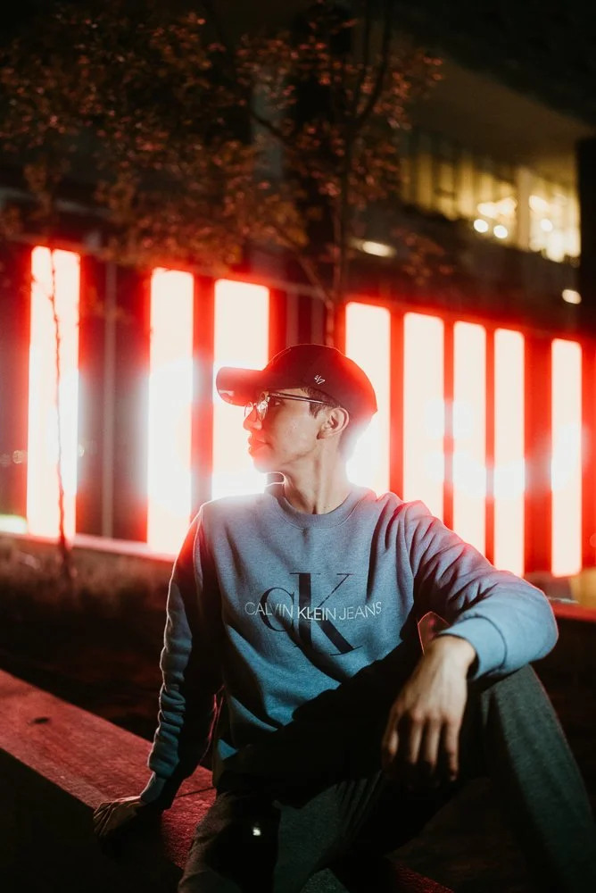 Person wearing a gray Calvin Klein sweatshirt and cap sitting outdoors at night, illuminated by vertical red and white neon lights in the background.