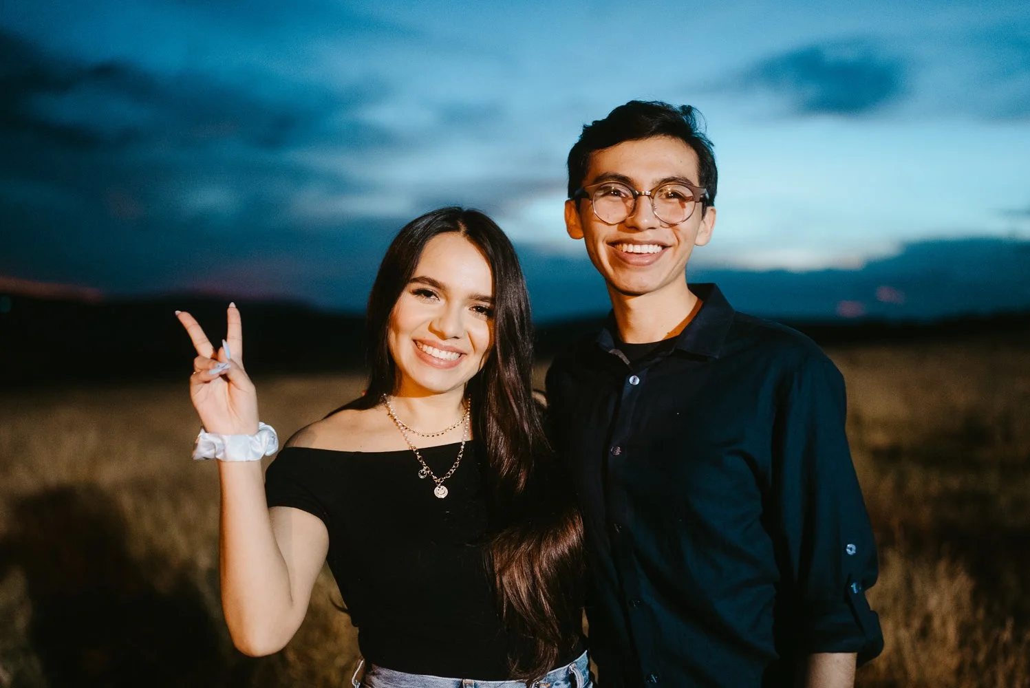 Two smiling teenagers, a girl and a boy, standing outdoors at dusk, with the sky darkening behind them. The girl has long dark hair, is wearing an off-shoulder black top, and makes a peace sign with her right hand. The boy has short dark hair, wears 