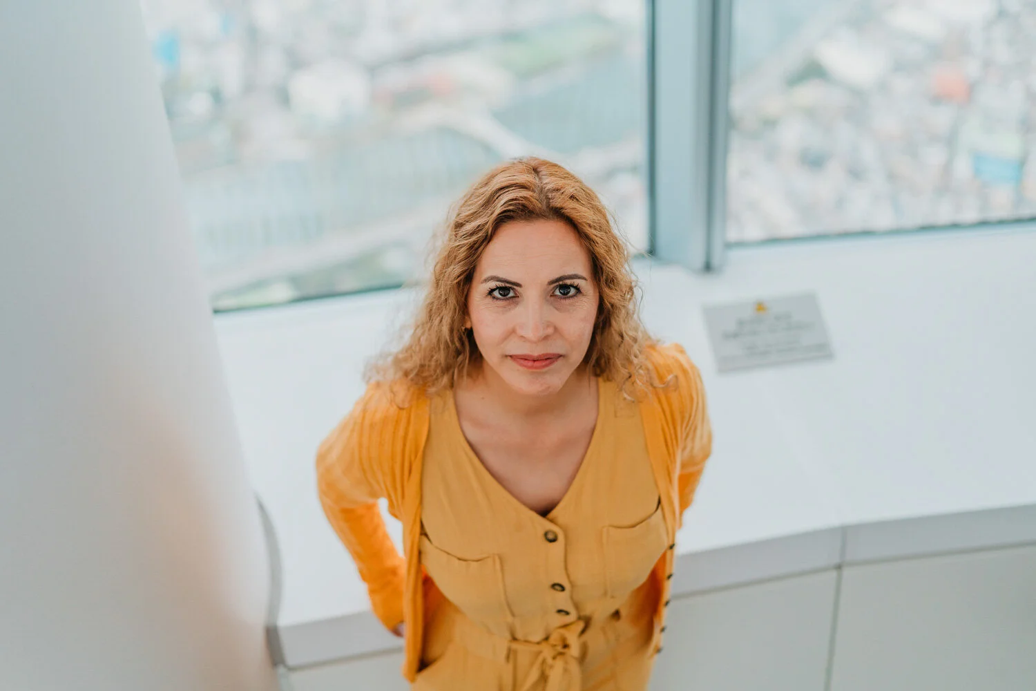 A woman with curly blonde hair wearing a yellow dress and orange cardigan, standing in front of large windows.