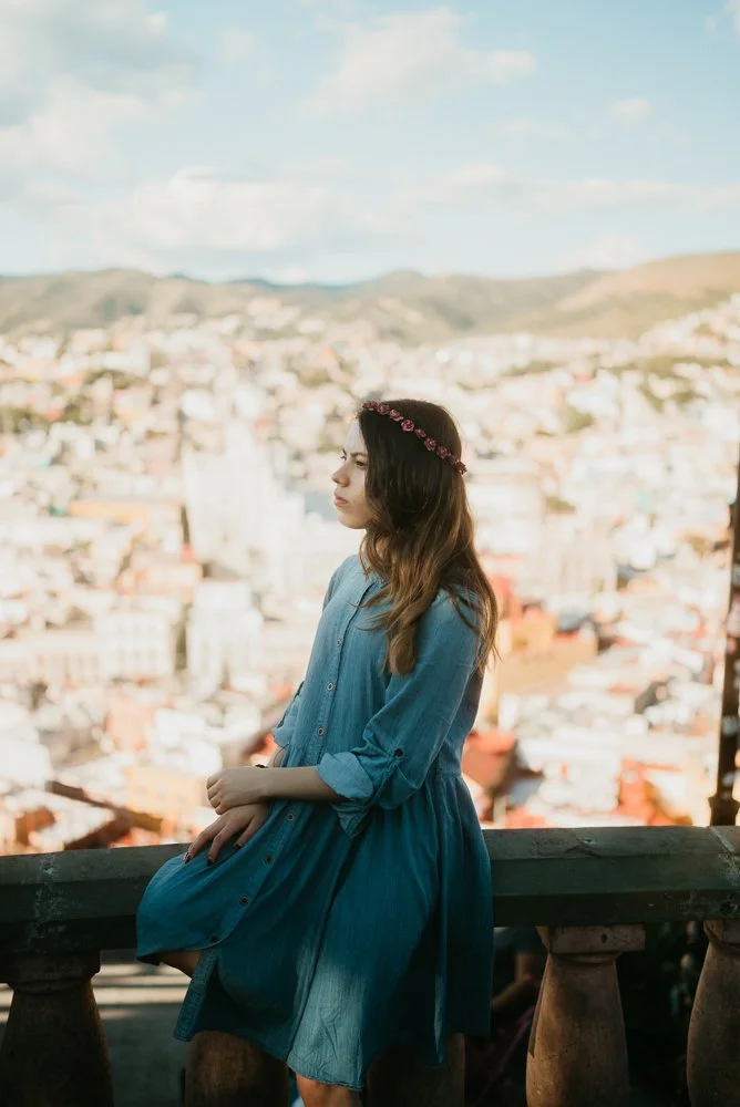 Young woman with long brown hair wearing a blue dress and a floral headband, sitting on a stone railing overlooking a cityscape with hills in the background.