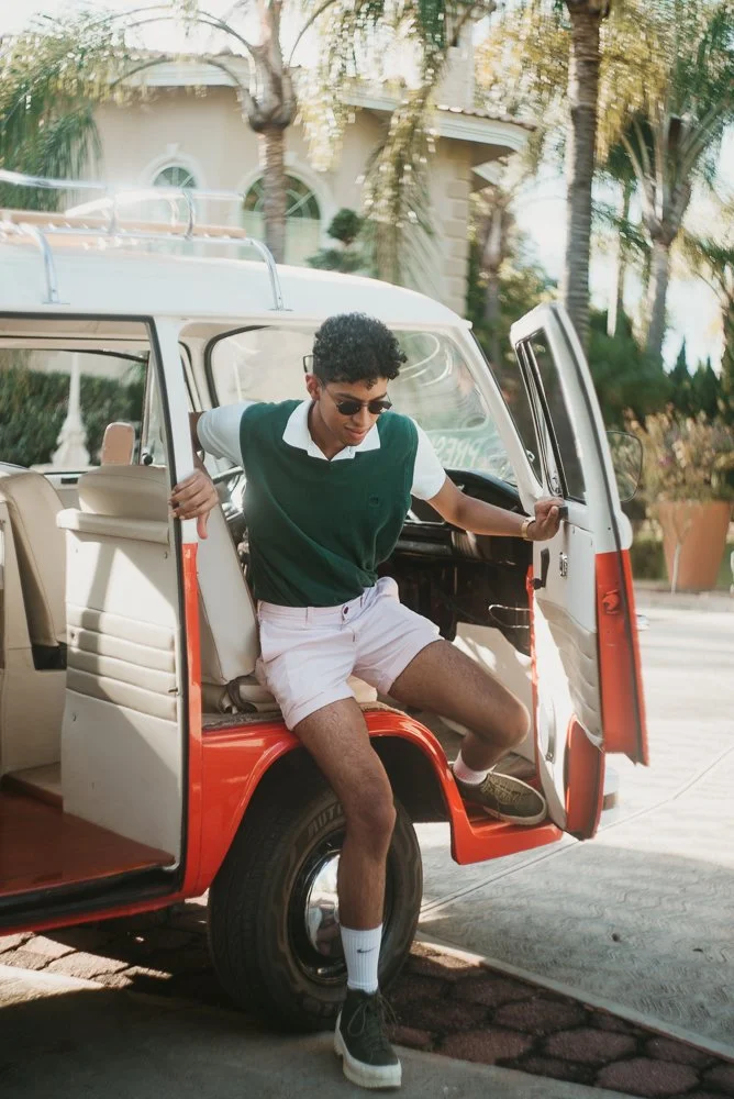 A young man with curly hair, sunglasses, a white shirt, green vest, and shorts is stepping out of a vintage red and white van parked on a residential street with trees and a house in the background.