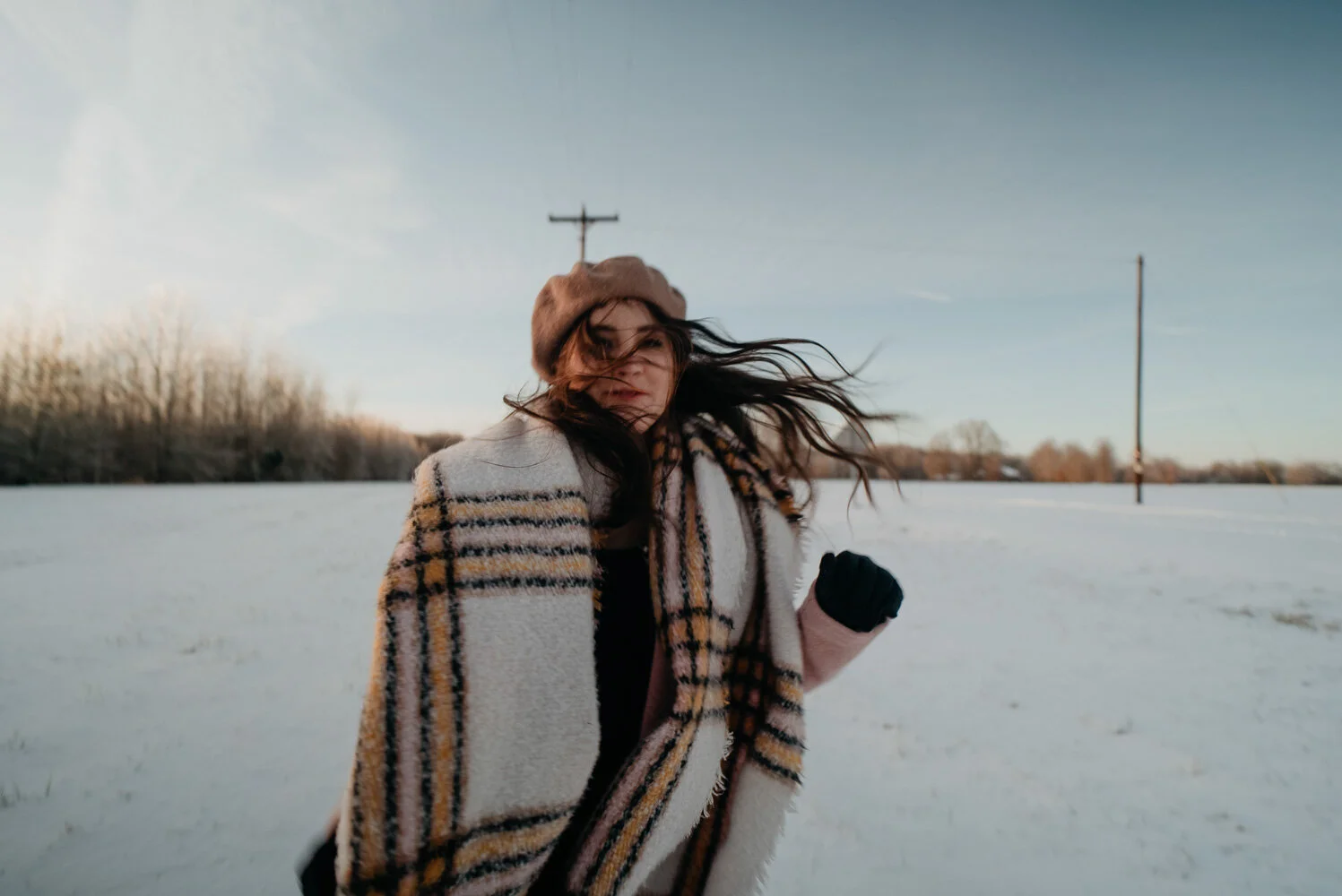 A woman standing outdoors in a snowy field, wearing a plaid coat, a tan beret, and a black glove, with her hair blowing in the wind during daytime.