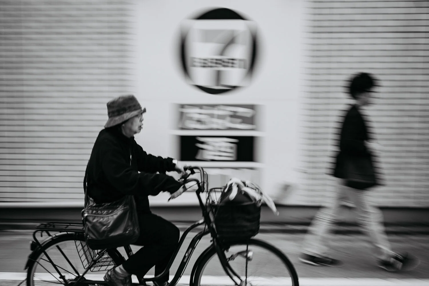 A woman riding a bicycle with a carrier basket, moving past a person walking in front of a textured wall with a blurred background.