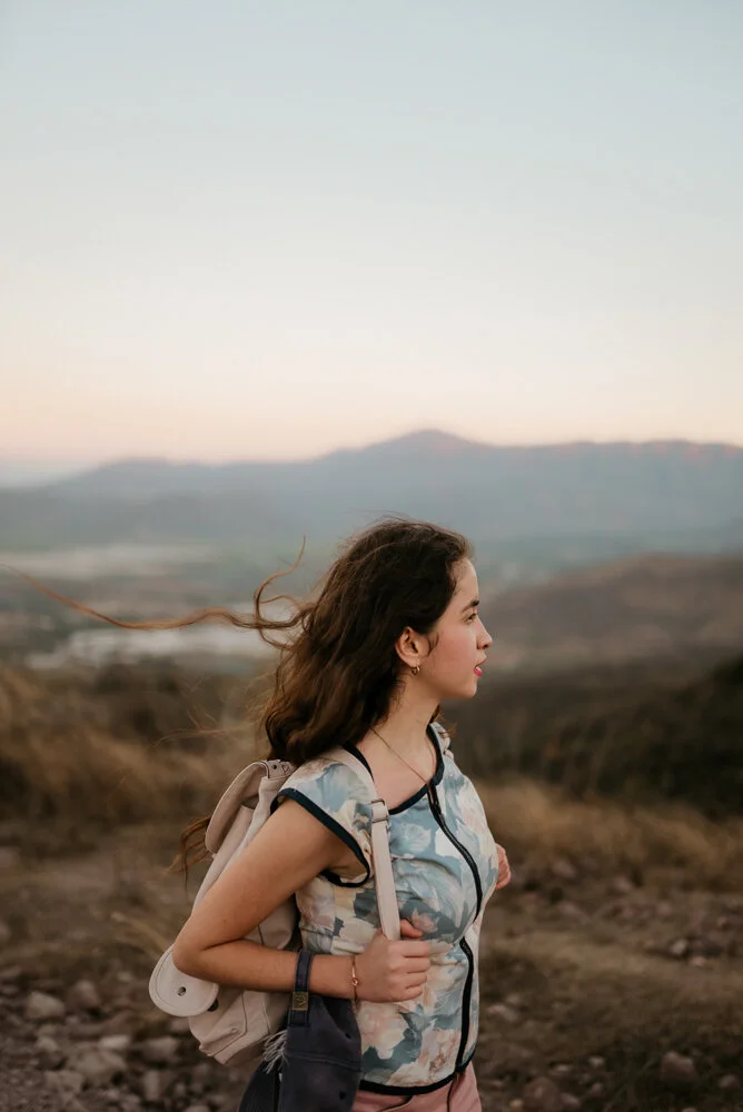 Young woman with dark brown hair in profile, wearing a patterned top and backpack, standing outdoors in a hilly landscape during sunset.