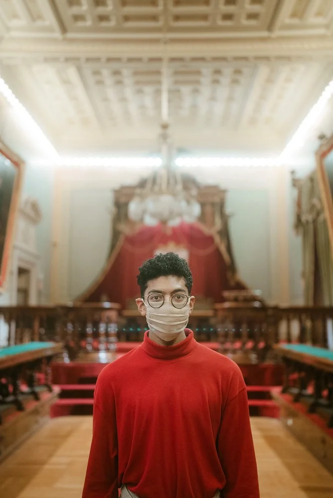 A young man wearing glasses and a face mask standing in an ornate, historic room with elaborate woodwork, high ceiling, chandelier, and red curtains.