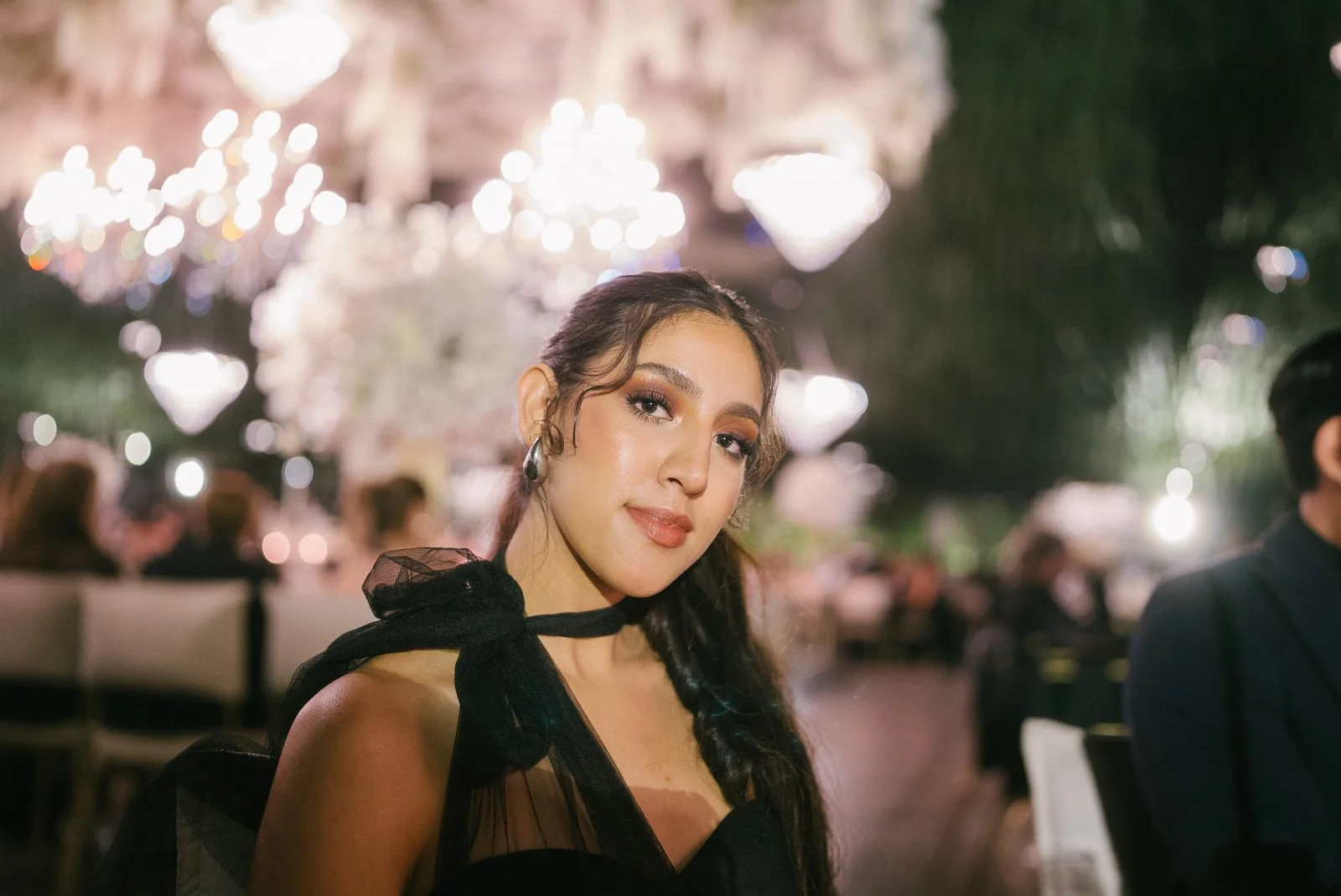 A young woman with long dark hair, wearing a black dress with sheer details and a bow, sitting at a formal event with chandeliers and blurred guests in the background.