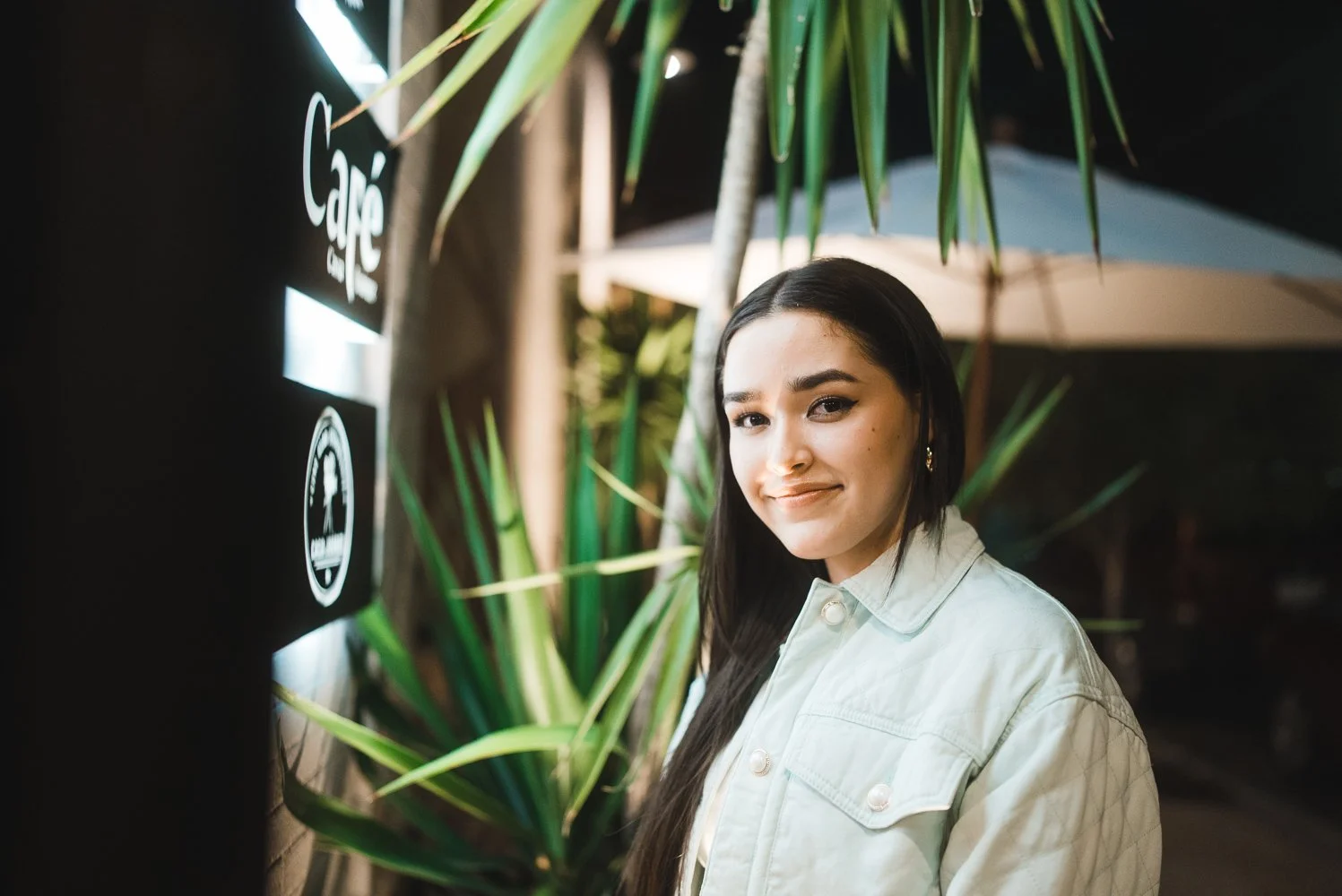 A young woman with long dark hair and earrings going to a cafe at night, standing near a glowing sign, with plants and a large umbrella in the background.