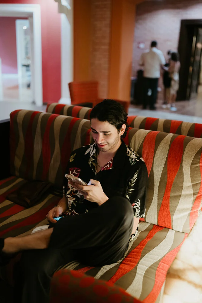 A young man with dark hair sitting on a multicolored striped couch, looking at his smartphone and smiling, in a warmly lit indoor setting.