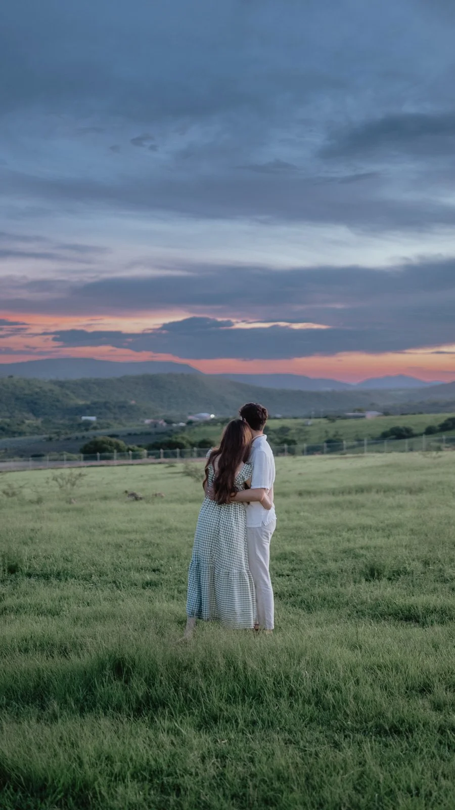 A couple standing in a grassy field at sunset, embracing and looking at the horizon with mountains in the distance.