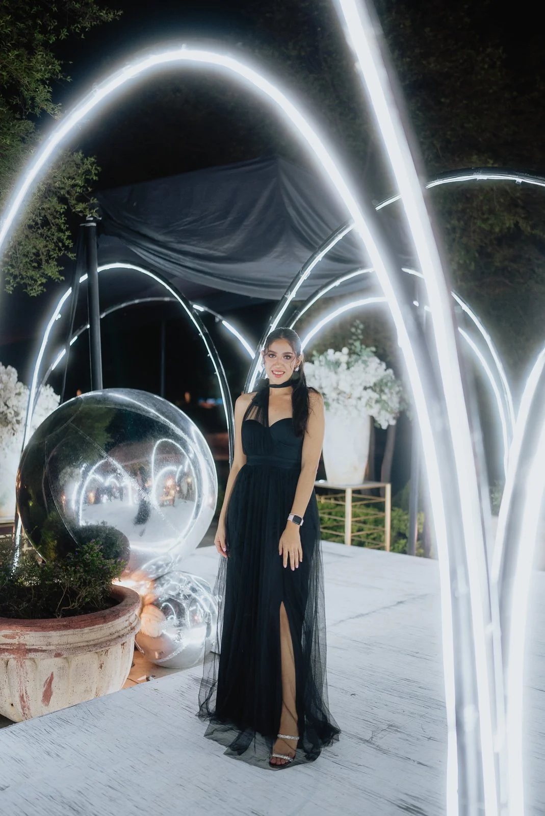A woman in a black gown standing under illuminated arches at night, with a glass globe and floral decorations nearby.