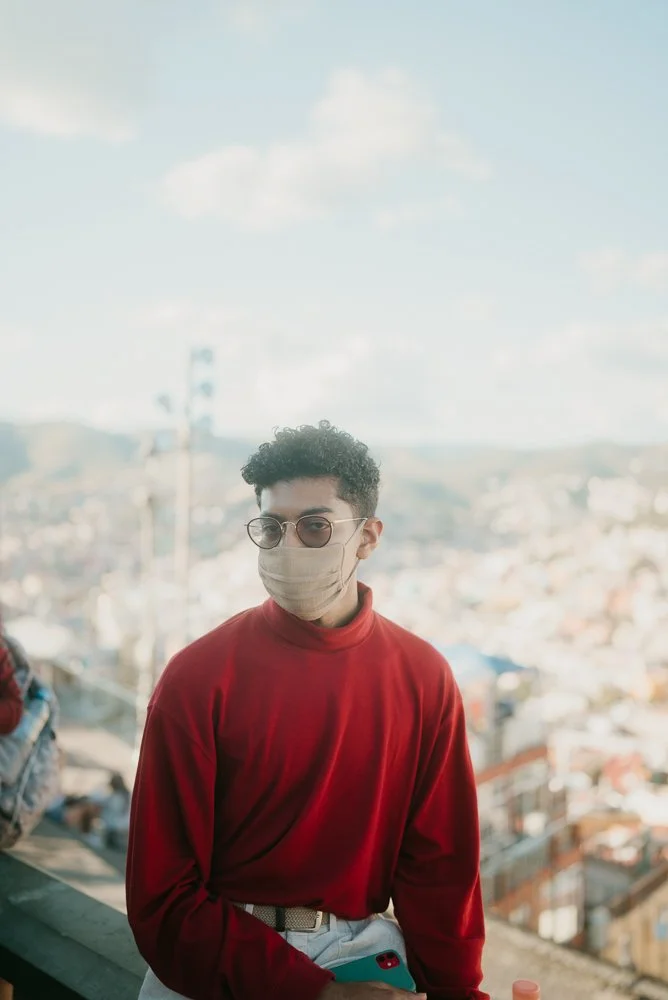 Young man with curly hair, wearing sunglasses, a face mask, and a red sweater, standing outdoors with a cityscape in the background under a partly cloudy sky.